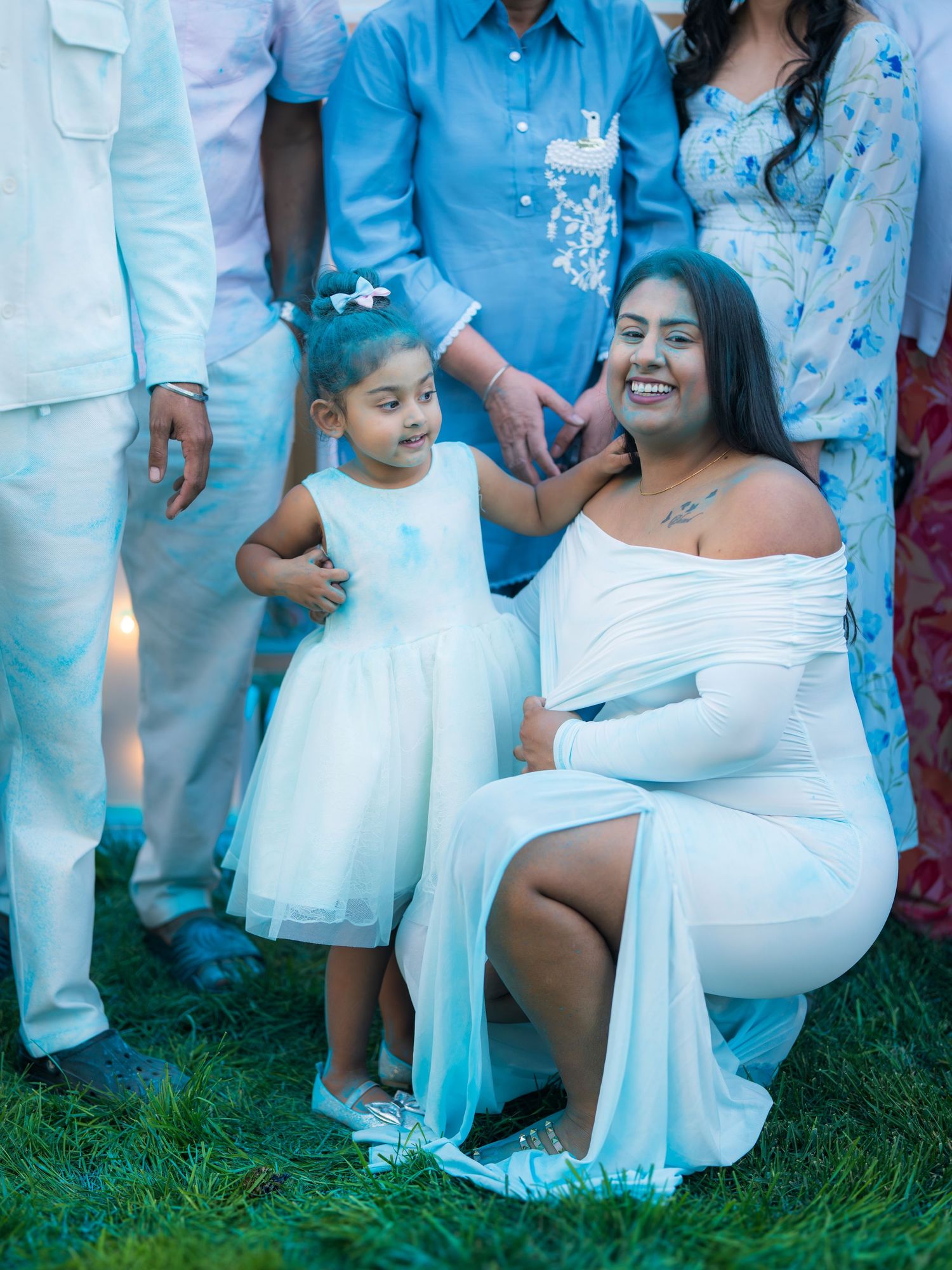 Pregnant woman in white dress crouching beside young girl in white dress at outdoor baby shower.