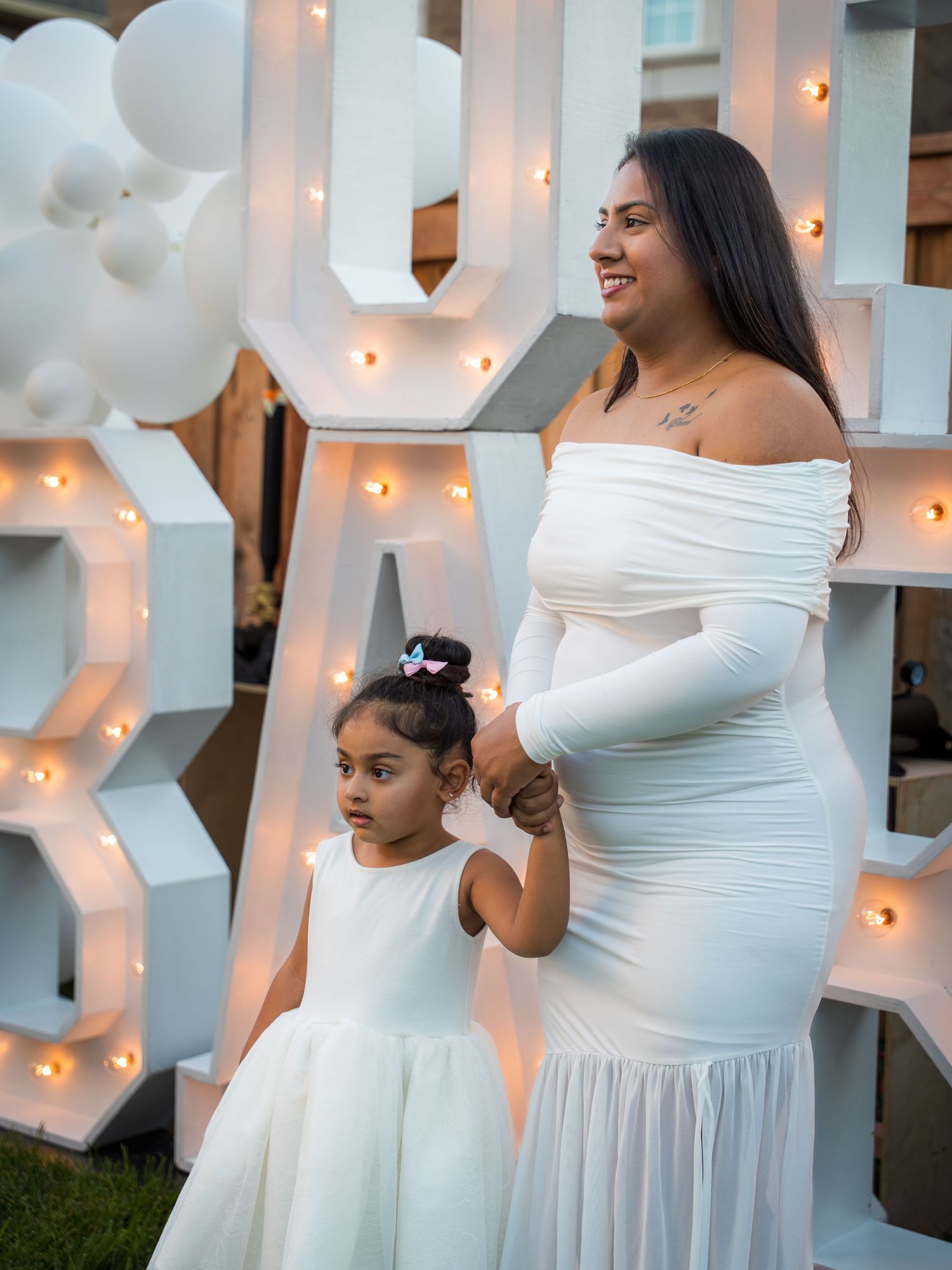 Pregnant woman in white off-shoulder dress holds hands with young daughter in front of OH BABY sign.