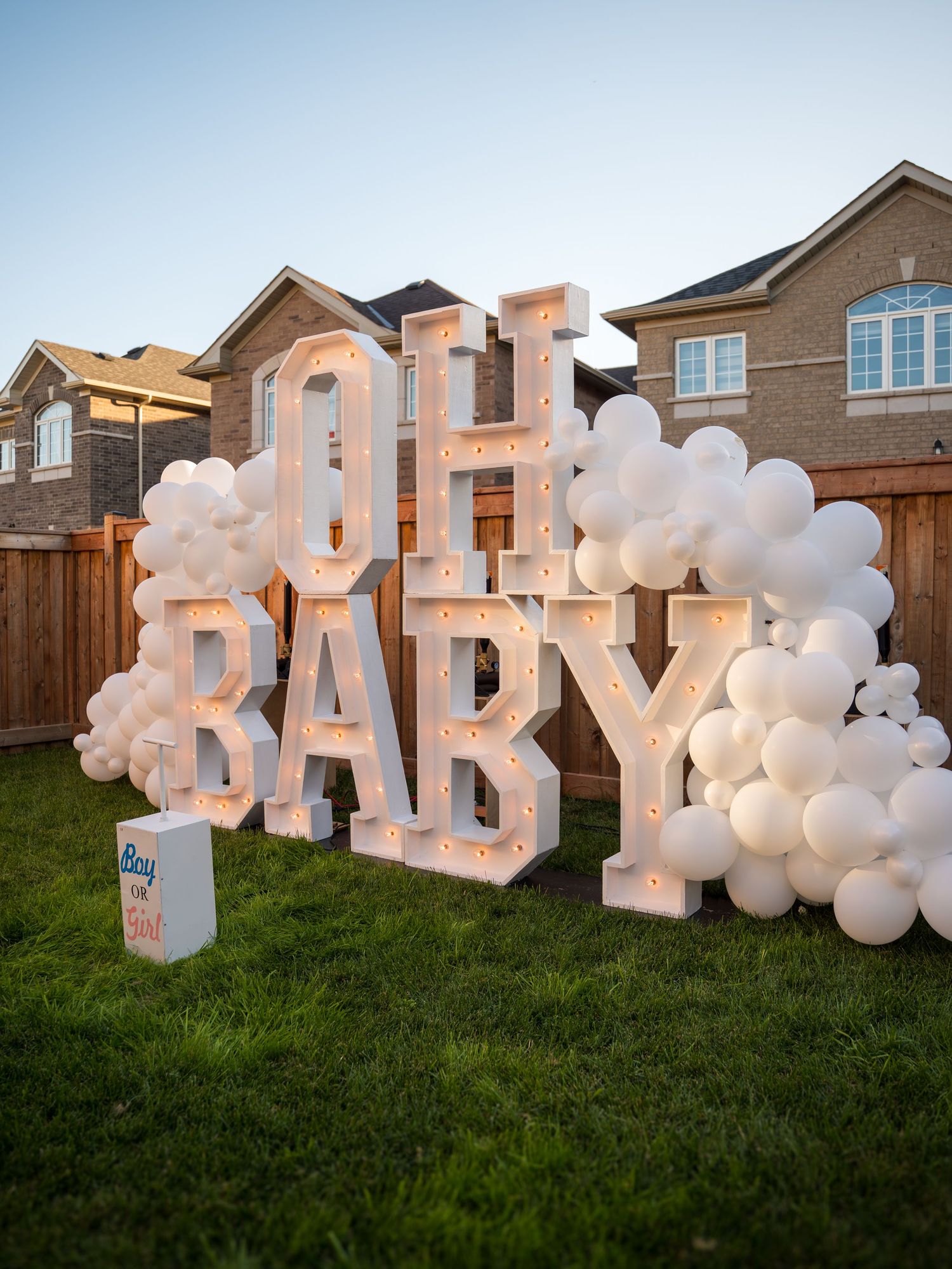Illuminated OH BABY marquee letter sign decorated with white balloon garland on green lawn at outdoor baby shower.