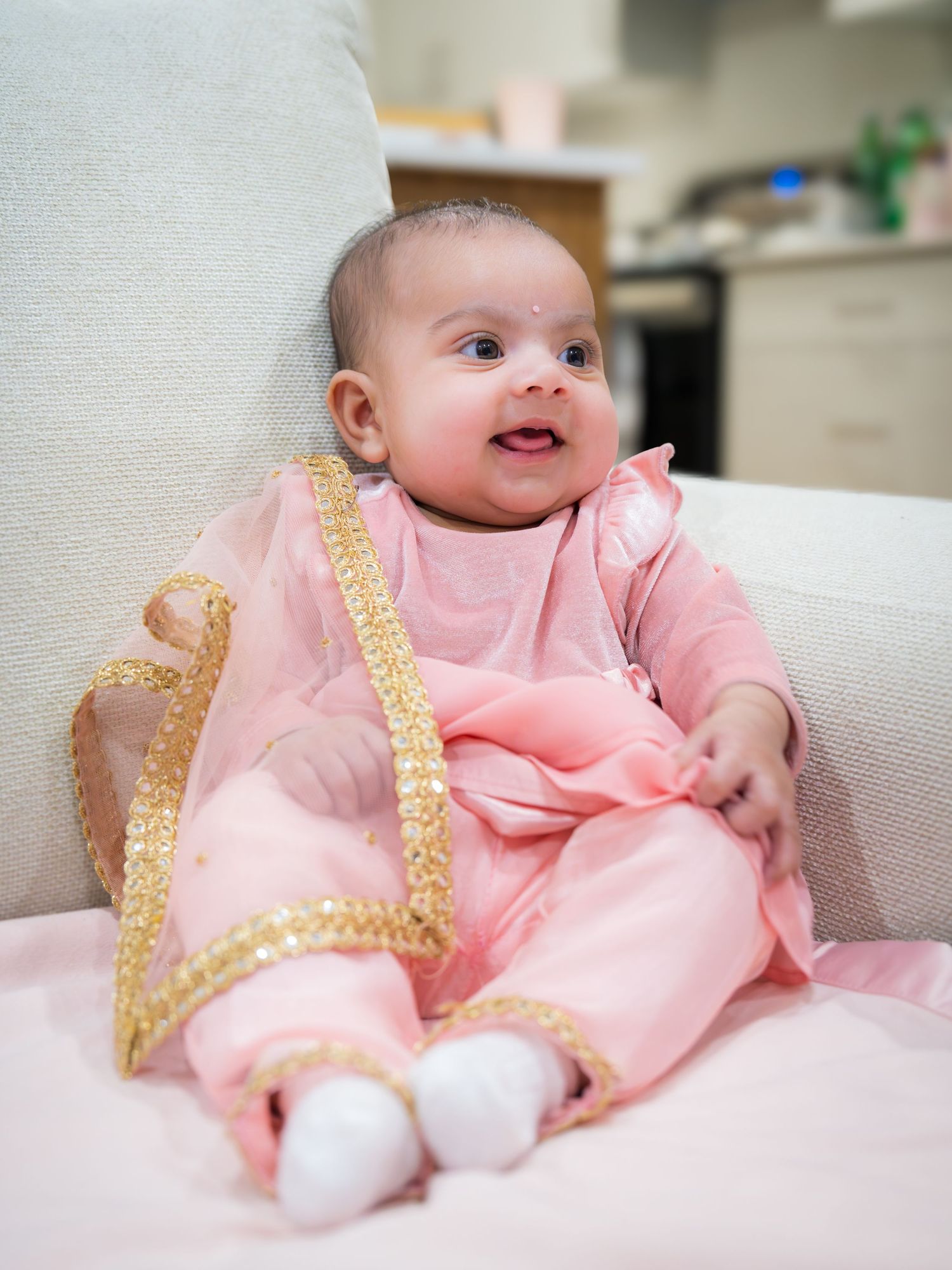 Cute baby girl smiling in pink and gold traditional Indian outfit seated on white couch at Lohri.