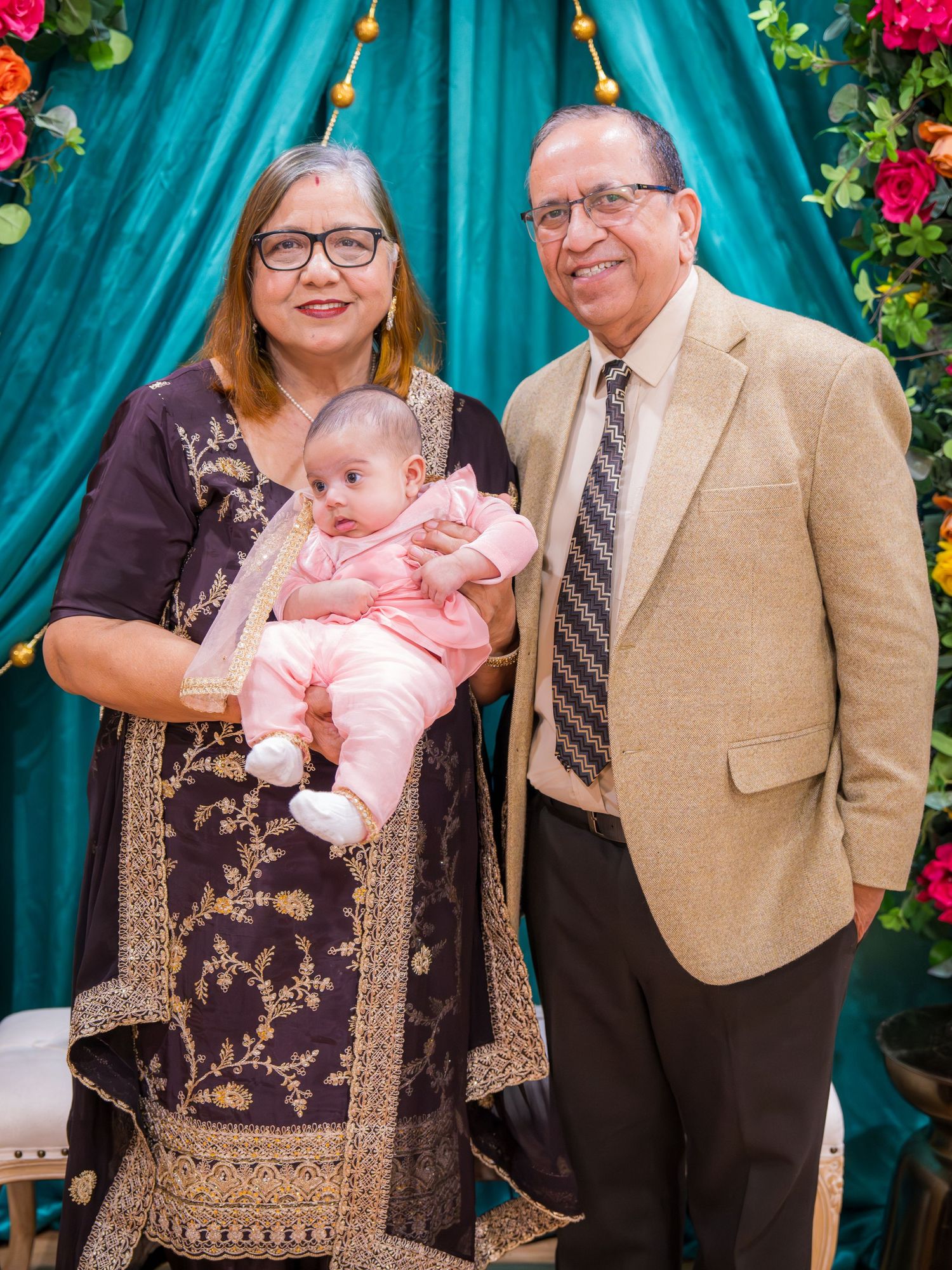 Grandparents holding infant baby dressed in pink at a colorful South Asian celebration event.