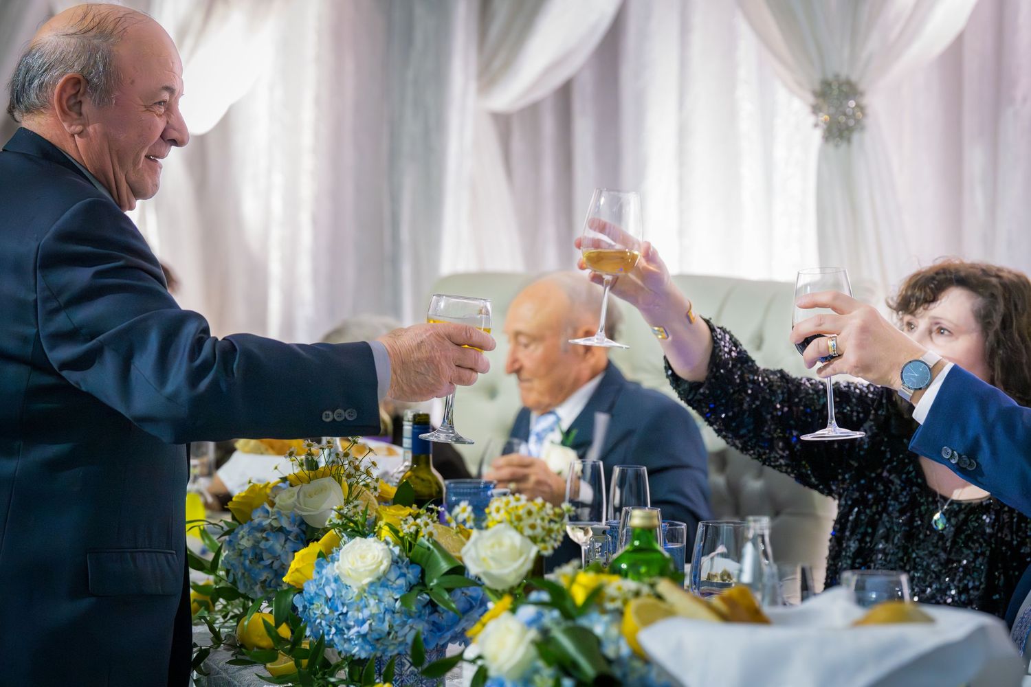 Elderly guests raising champagne glasses in a toast at a wedding reception with yellow and blue floral centerpieces.