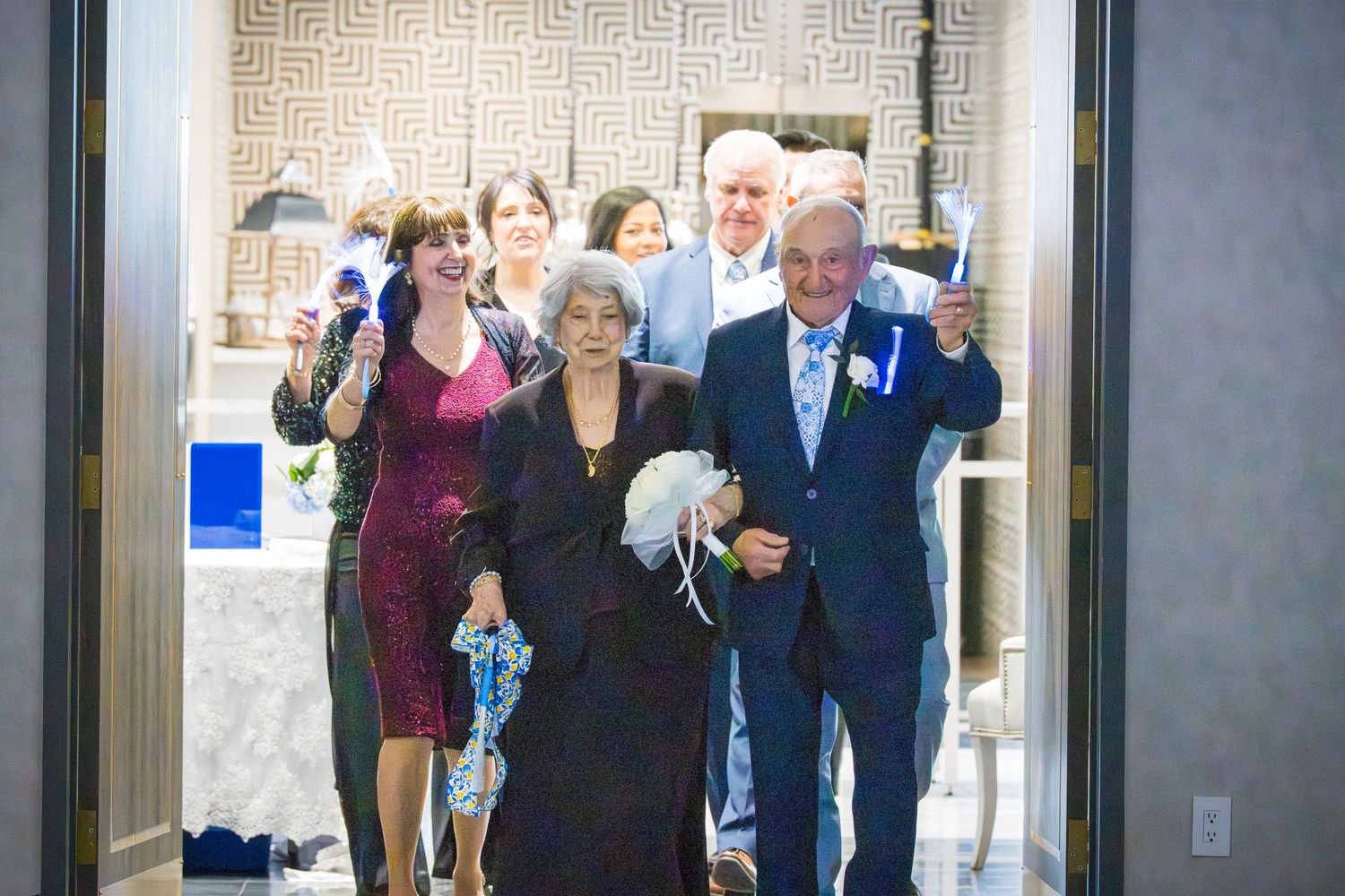Elderly couple making grand entrance at celebration venue with guests cheering and waving blue pompoms behind them.