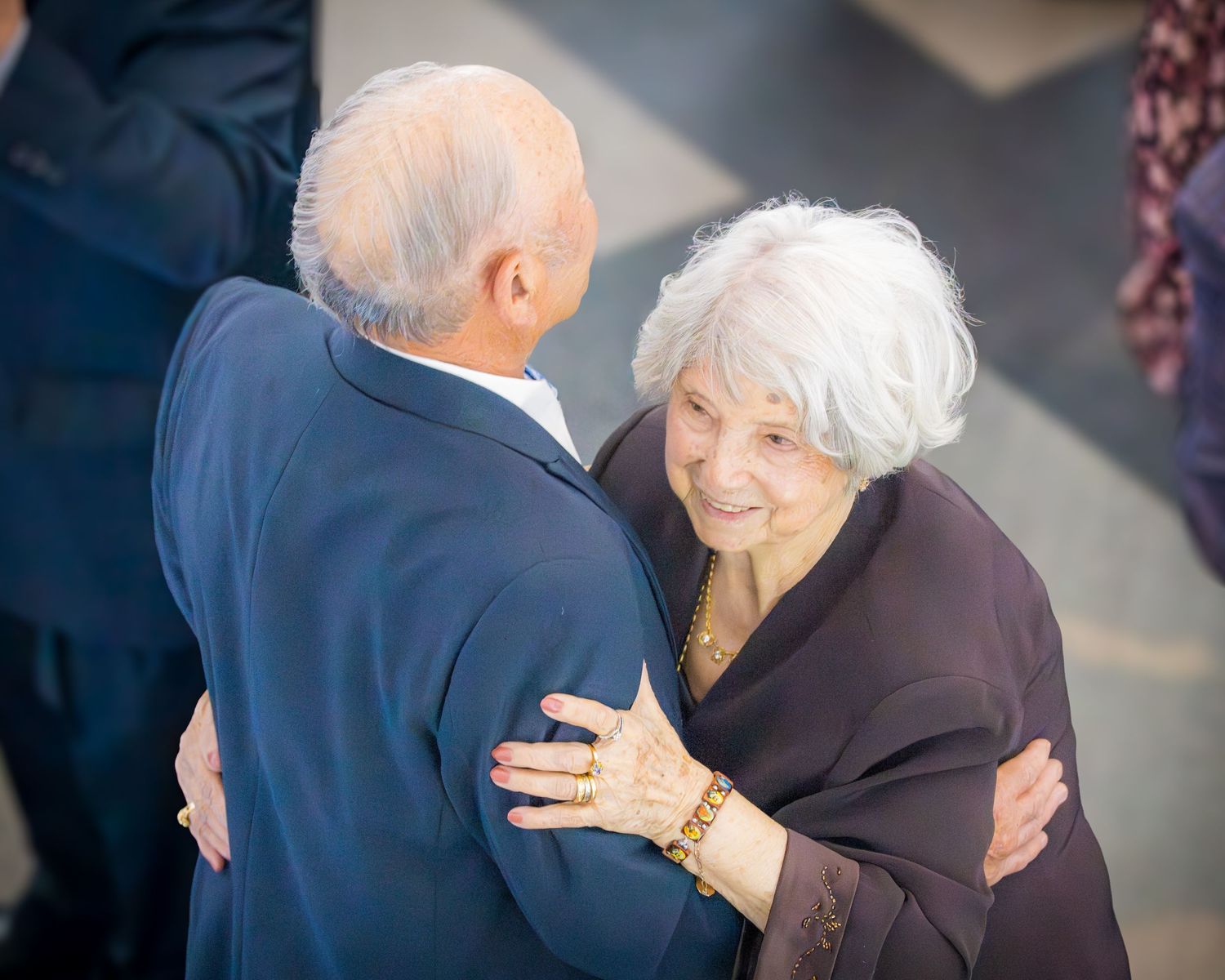 Elderly man and woman warmly embracing and smiling at each other outdoors at a formal celebration.
