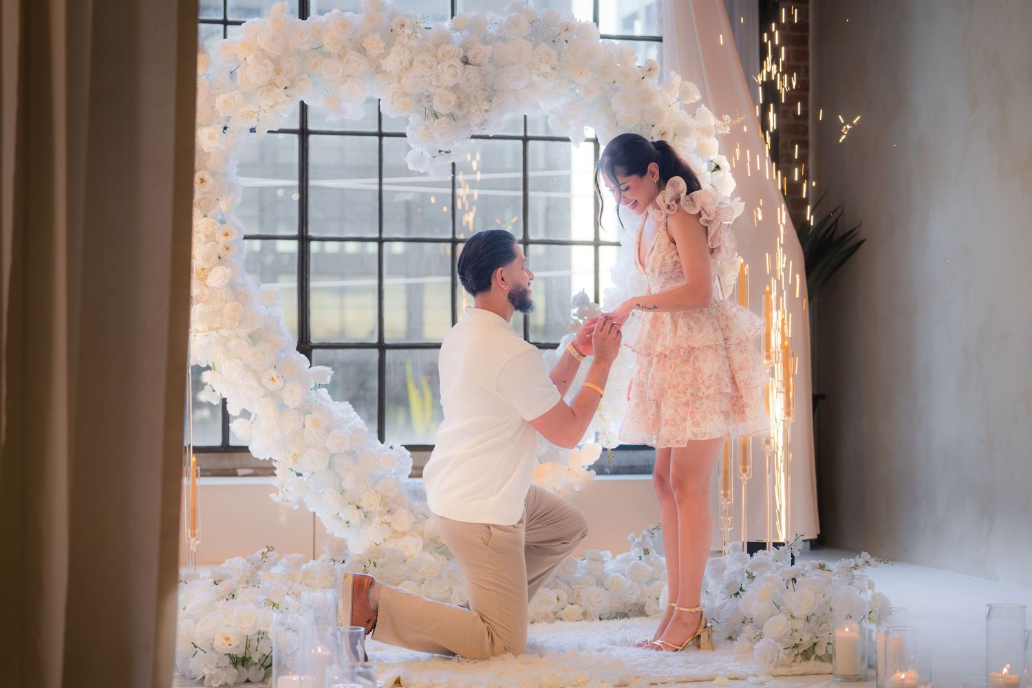 Man proposing on one knee to woman inside a white heart floral arch with candles.