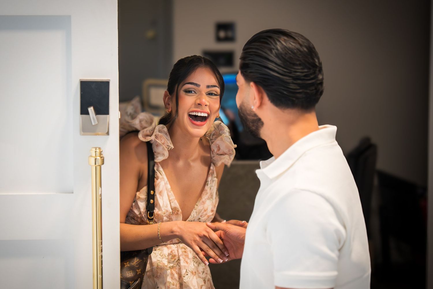A joyful couple sharing a happy moment at a doorway, woman laughing in a floral dress as man holds her hands.