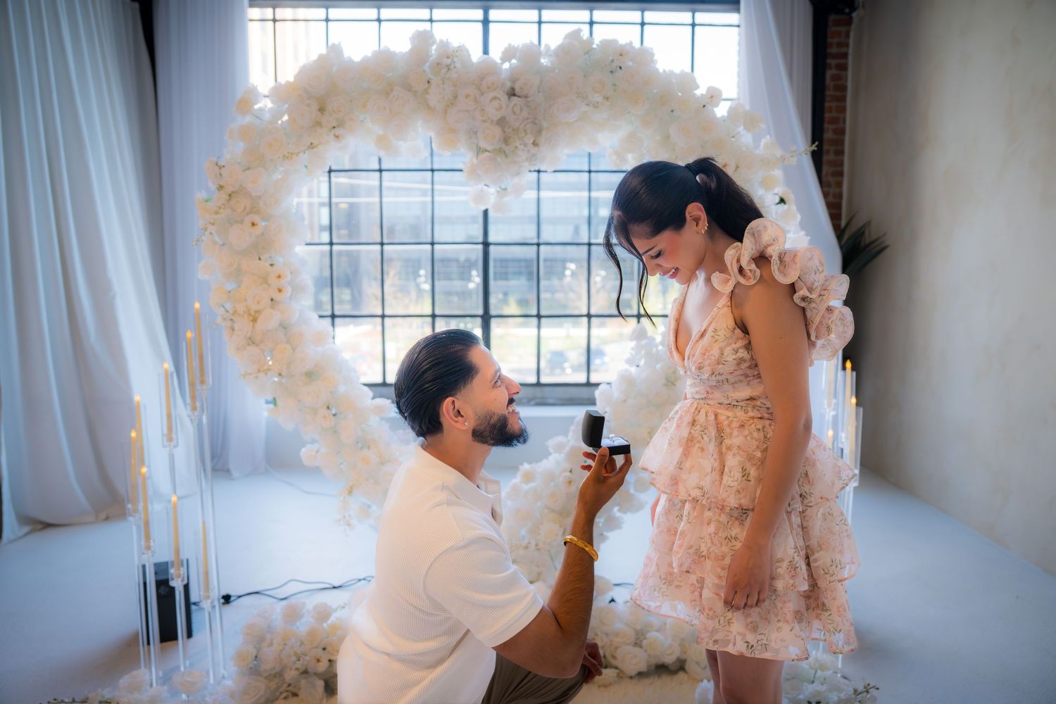 Man proposing with ring box to woman in front of large white floral heart arch.