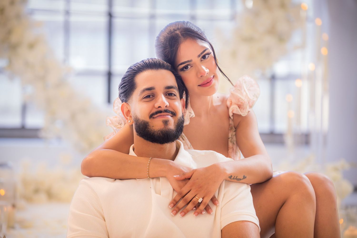 Couple posing together in white outfits surrounded by white floral decorations during an engagement photoshoot.