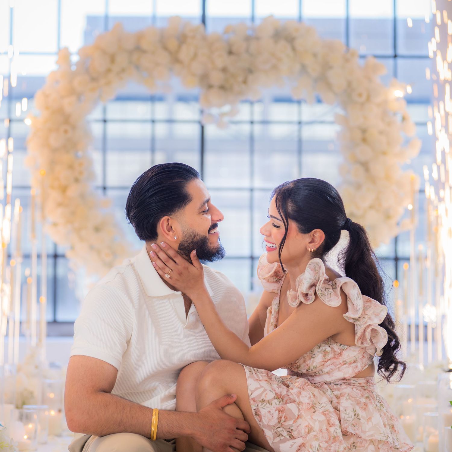 Smiling couple gazing at each other before illuminated white heart floral arch.