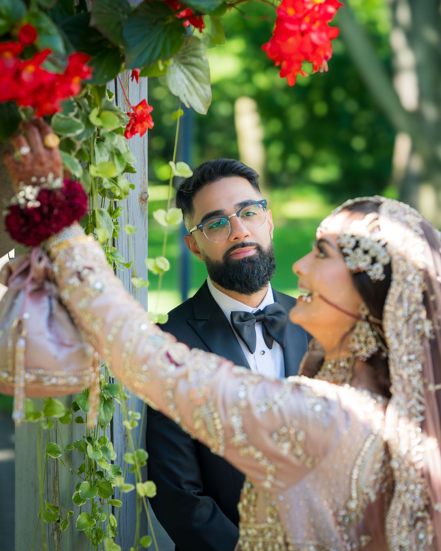A couple in wedding attire share a romantic moment in a garden setting with vibrant red flowers.