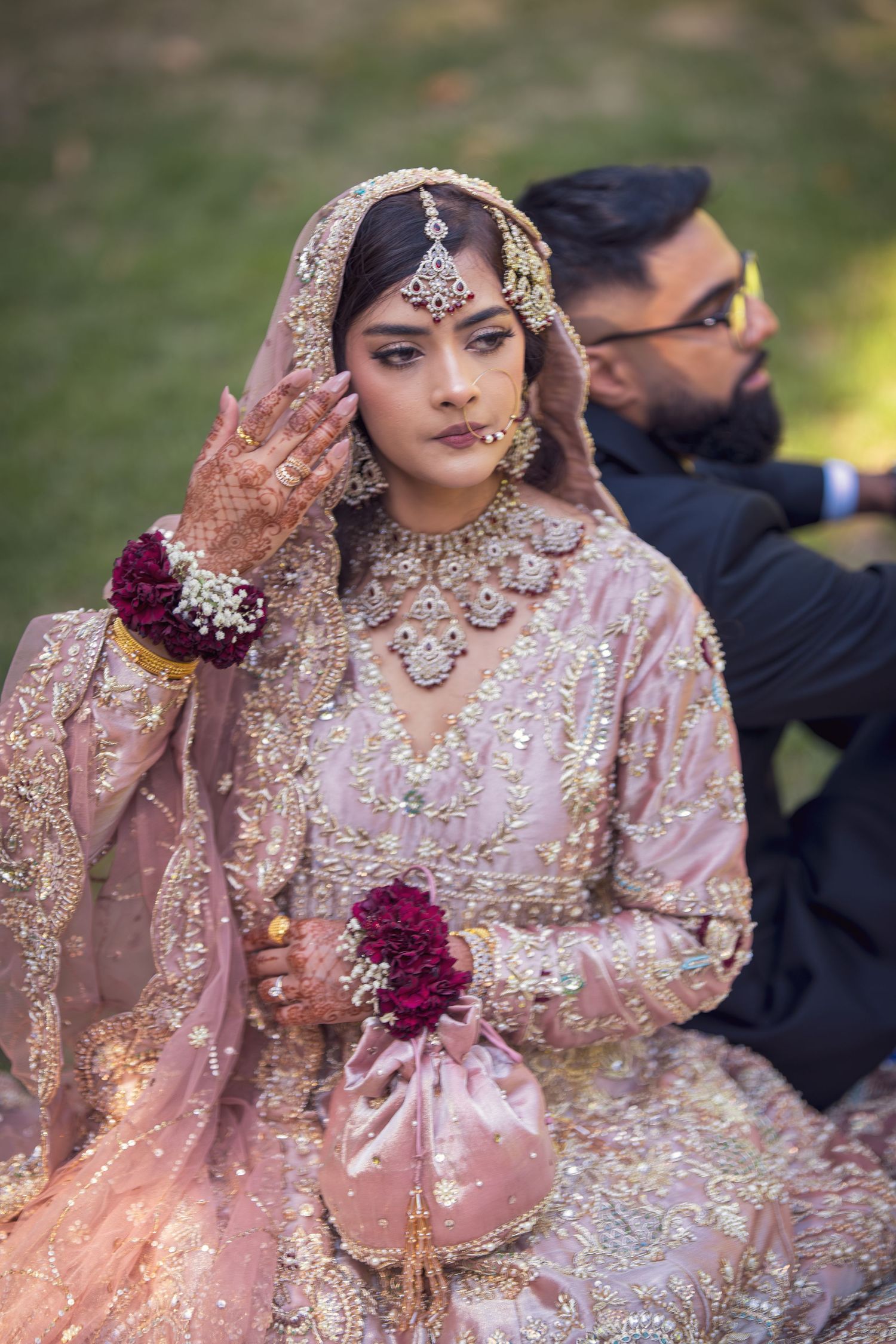 Bride in embellished pink lehenga with intricate jewelry and henna poses during outdoor wedding photoshoot.
