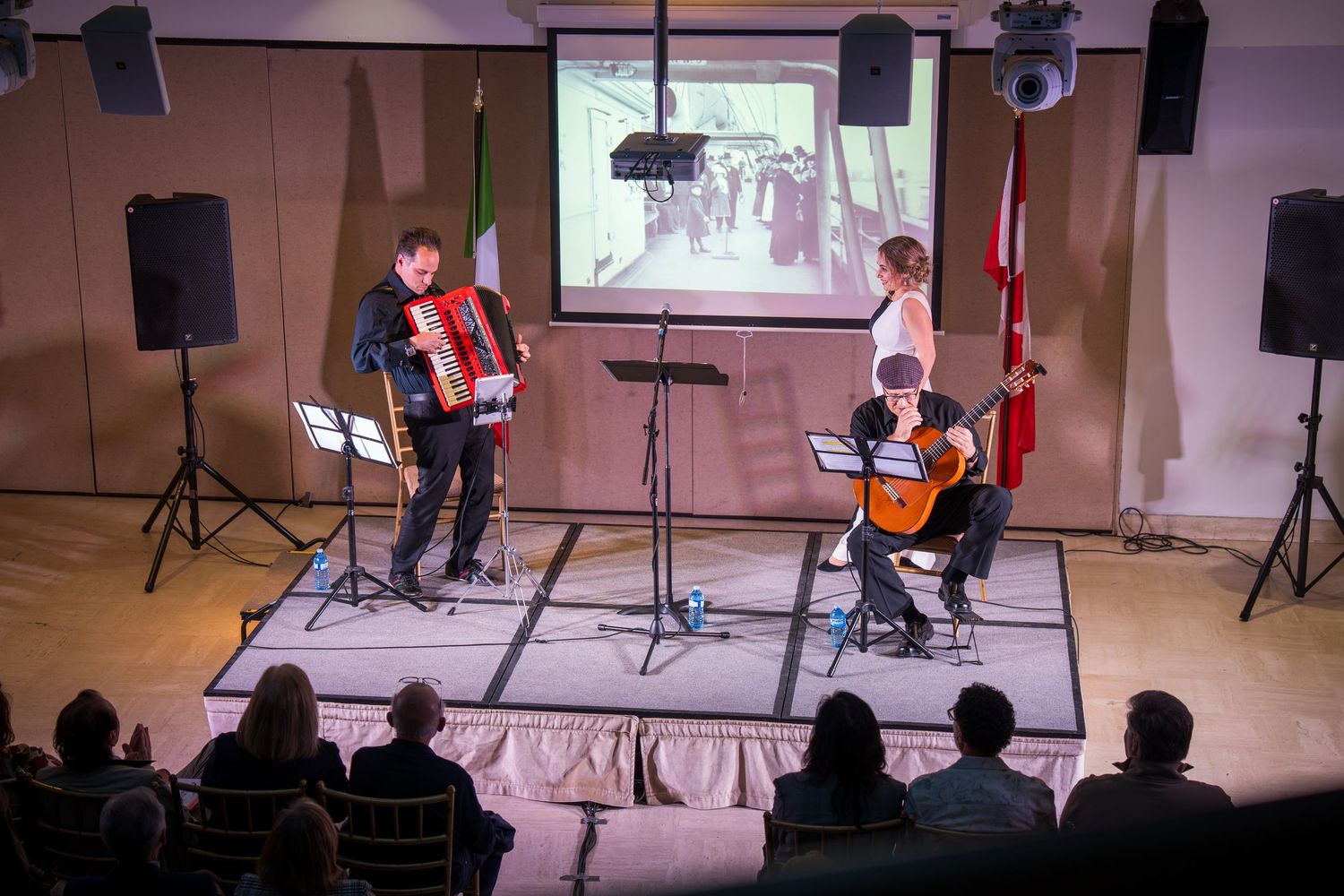 Musicians perform accordion and guitar on stage with audience watching and a projected image displayed behind them.