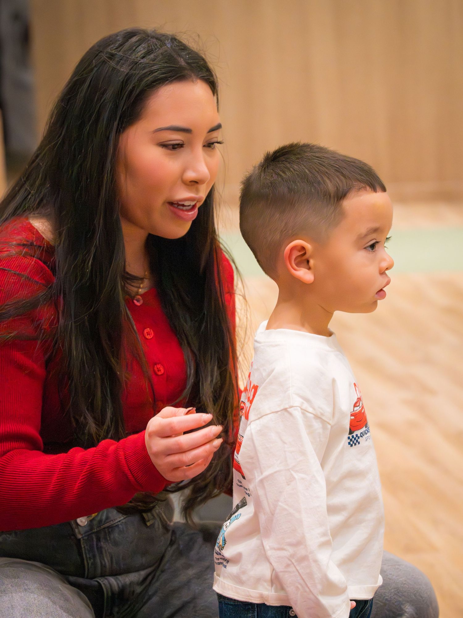 Young woman in red sweater kneeling beside a toddler boy at an indoor event.
