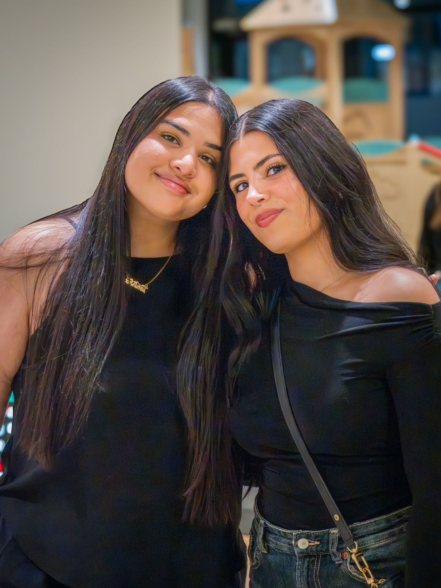 Two young women with long dark hair wearing black outfits smile together at an indoor event.