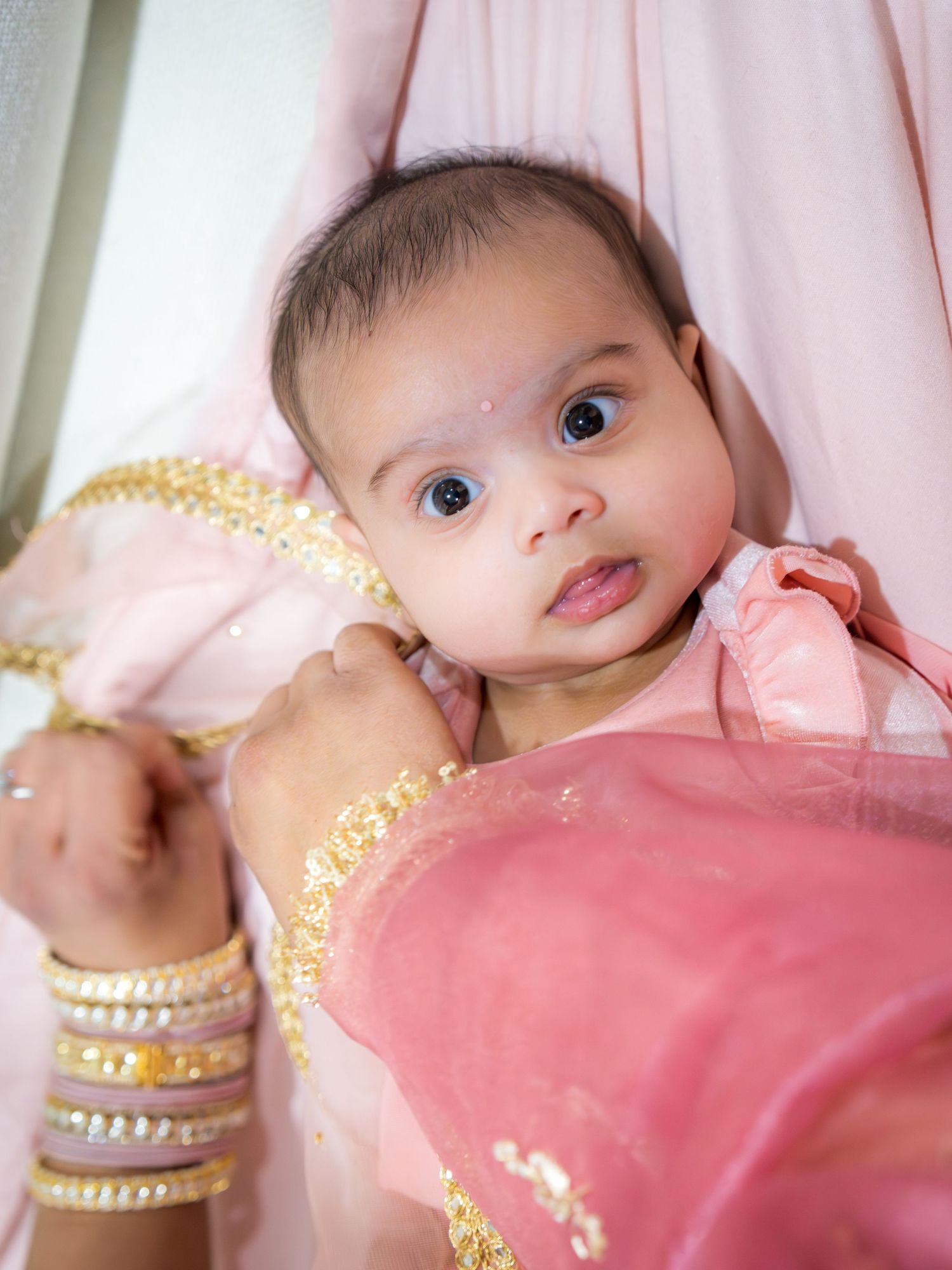 Adorable baby in pink embroidered outfit with gold trim at an Indian celebration ceremony.