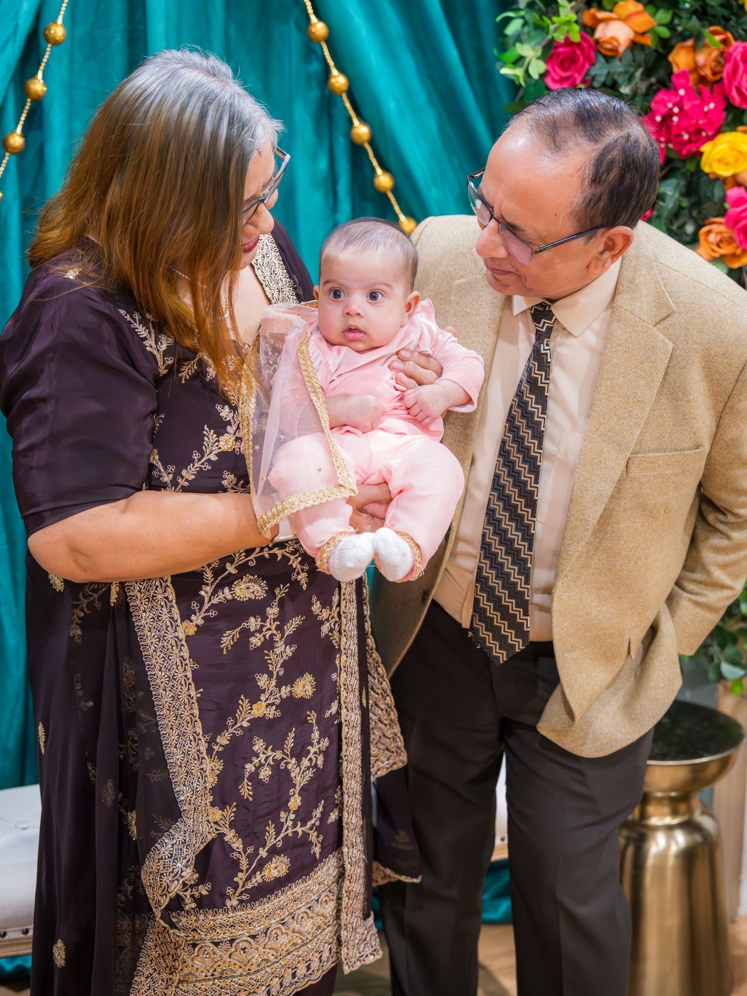 Grandparents in traditional South Asian attire lovingly hold pink-dressed newborn at family event.