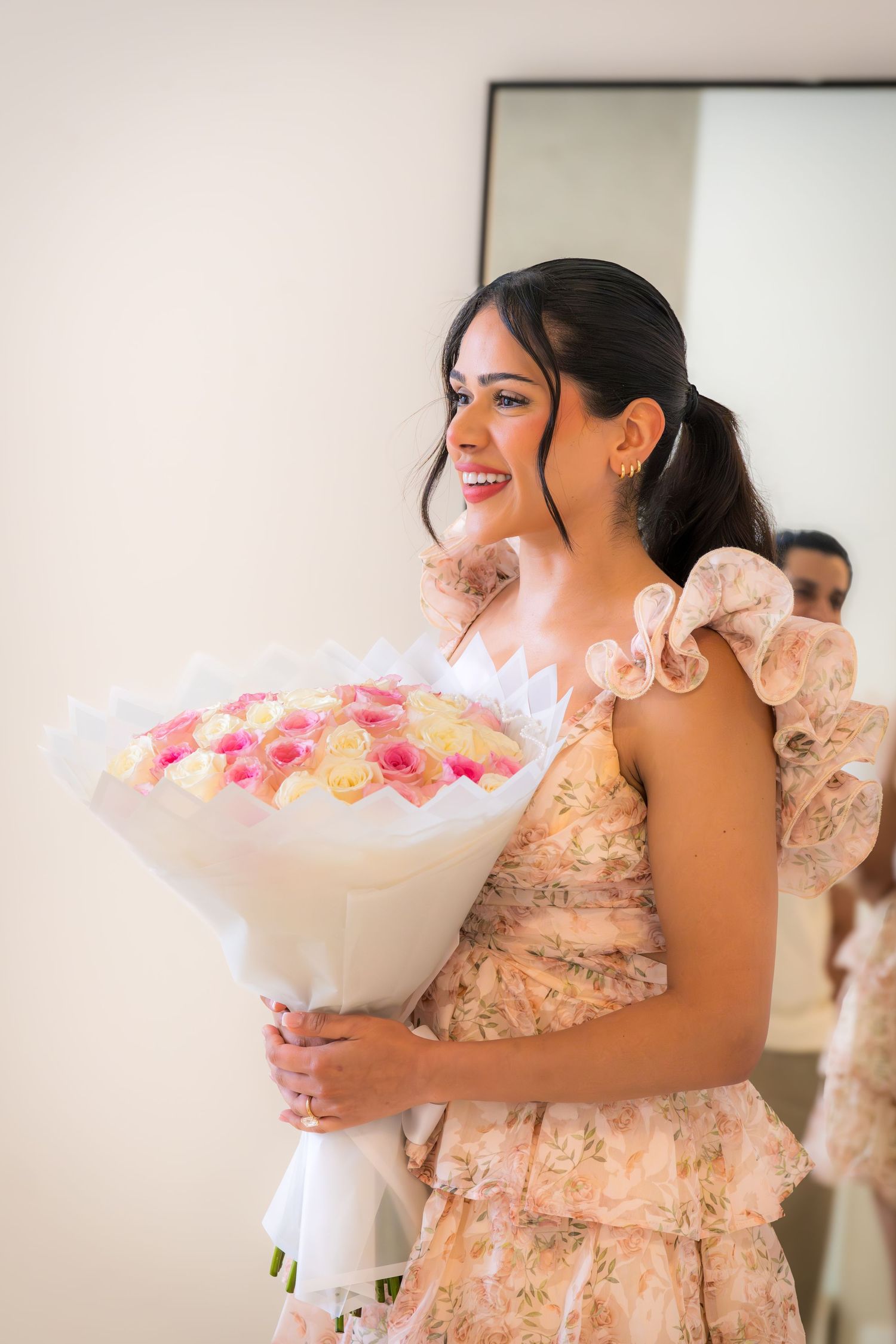 Smiling bride holds large pink and white rose bouquet wearing floral ruffled dress.
