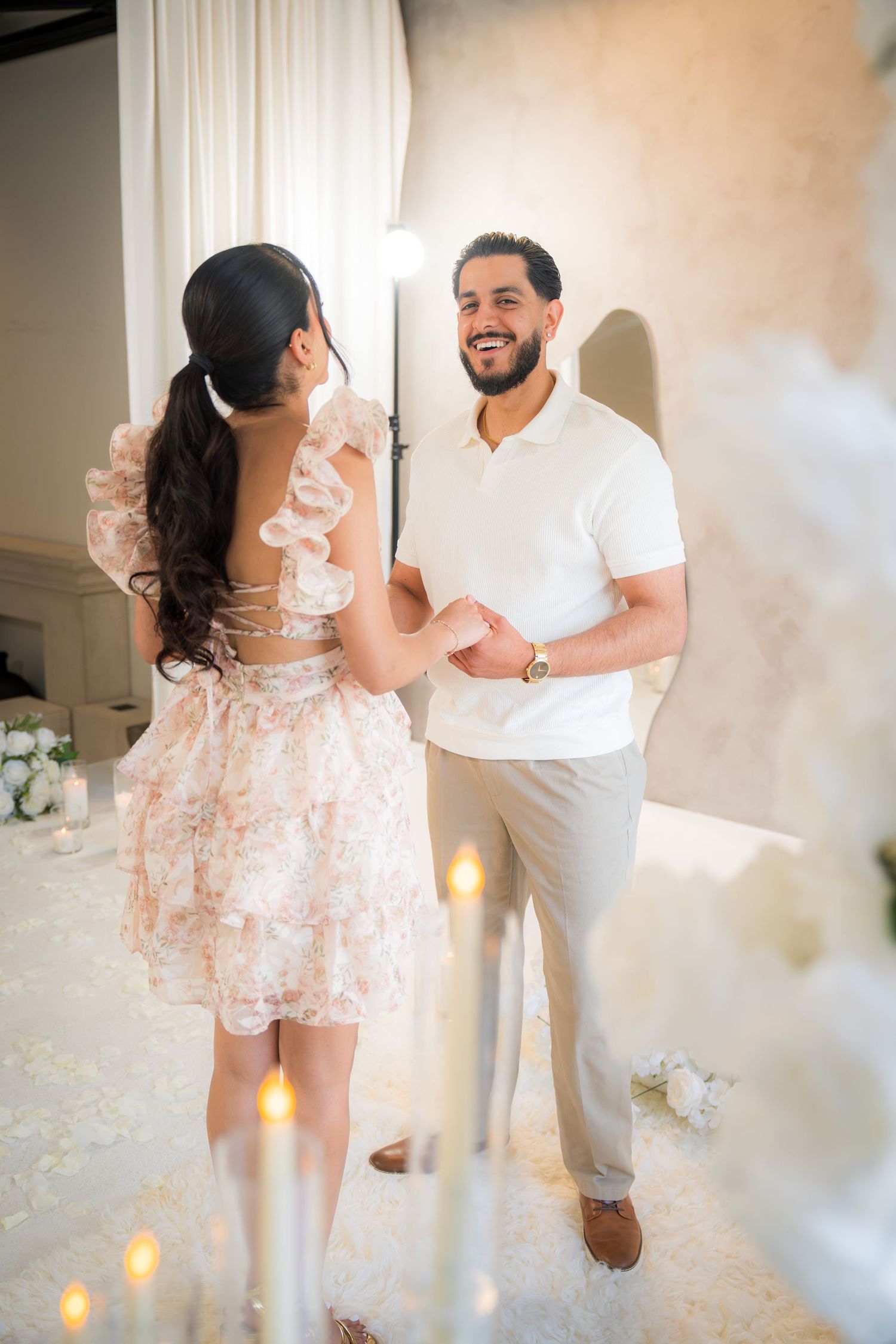 Couple holding hands and smiling during a romantic proposal setup with candles and white floral decorations.