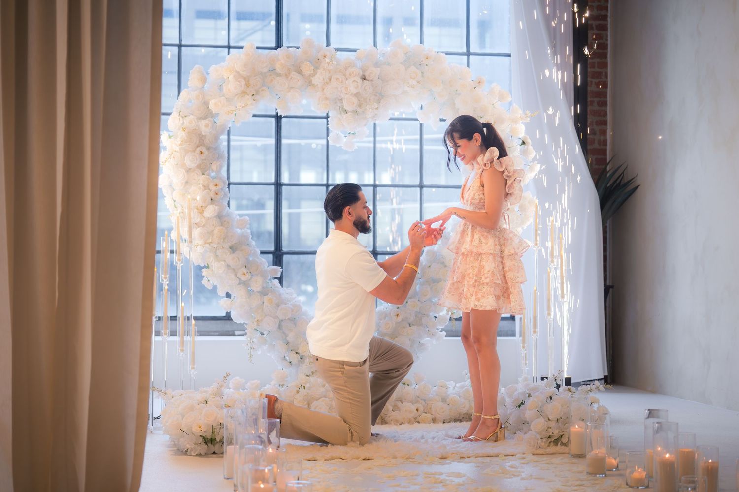 Man on one knee proposing to his partner in front of a large white floral heart arch with candles nearby.