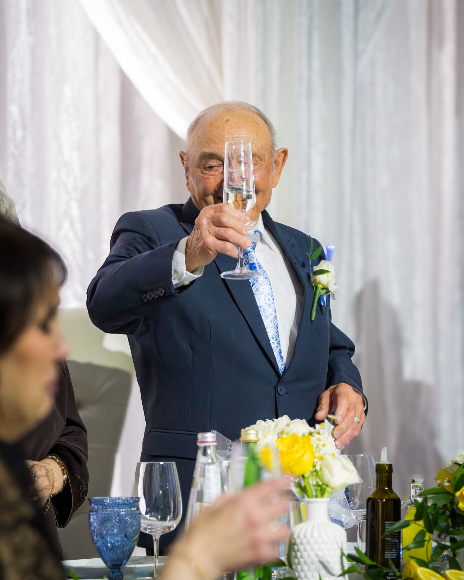 Elderly man in navy suit raising champagne glass for a toast at a wedding reception.