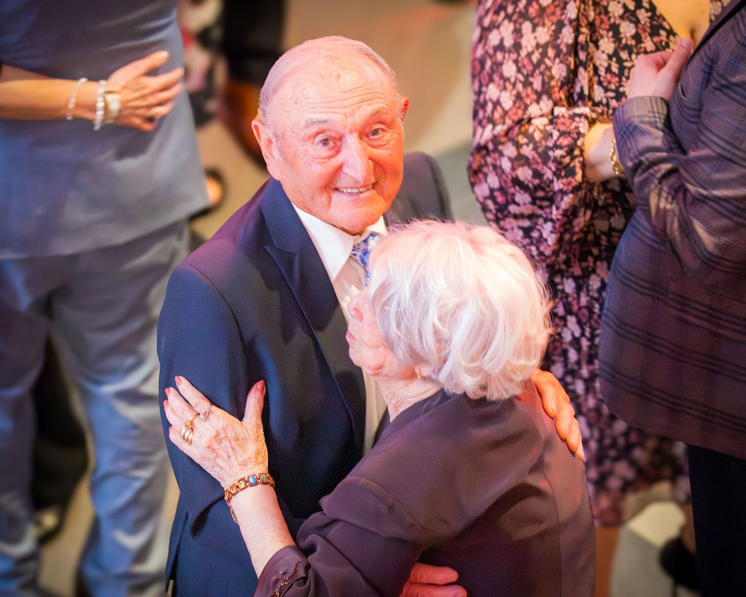 Elderly man in blue suit warmly embracing a white-haired woman at a formal celebratory gathering.