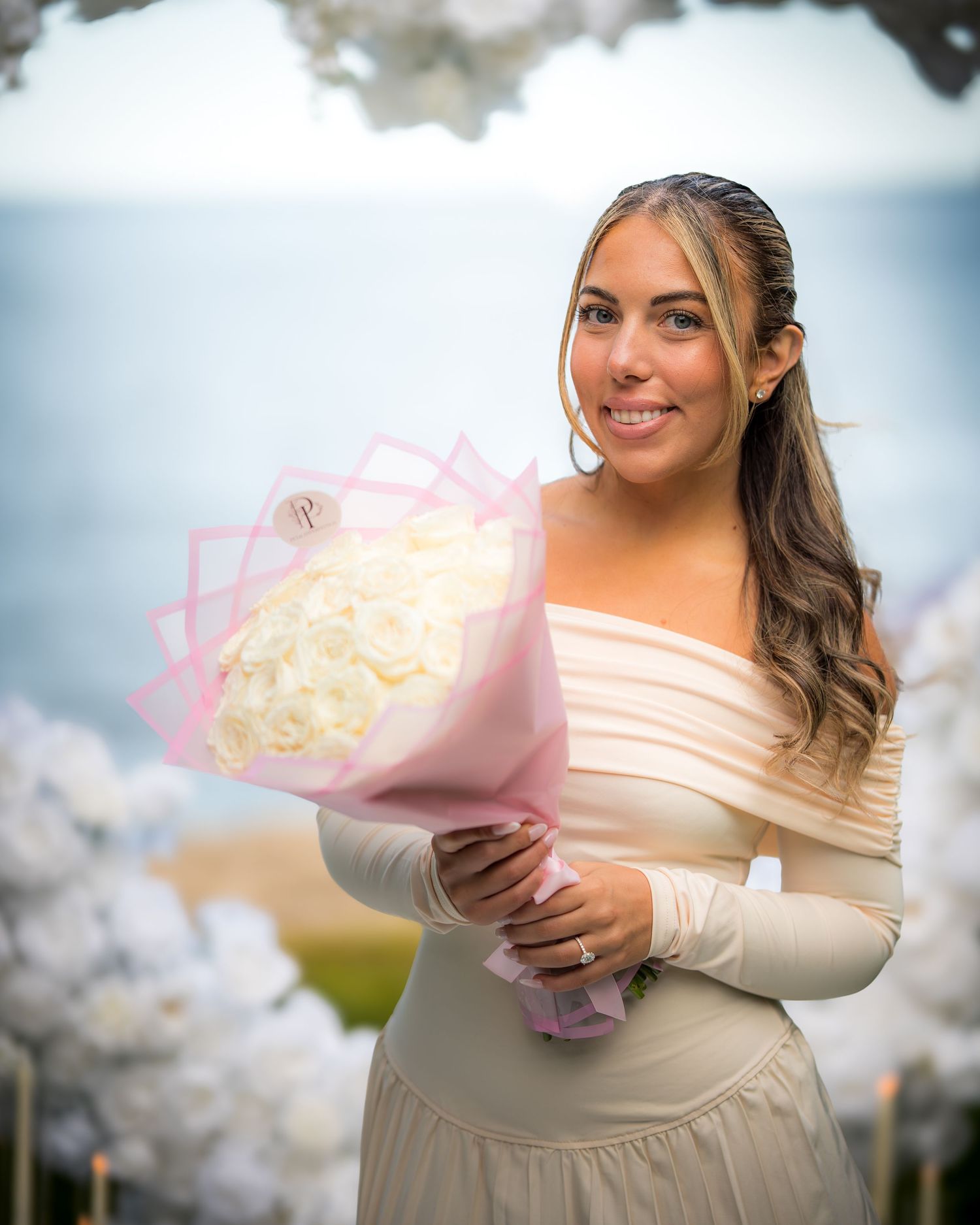 A person in a cream dress holds a pink-wrapped bouquet of white flowers against a cloudy sky background.
