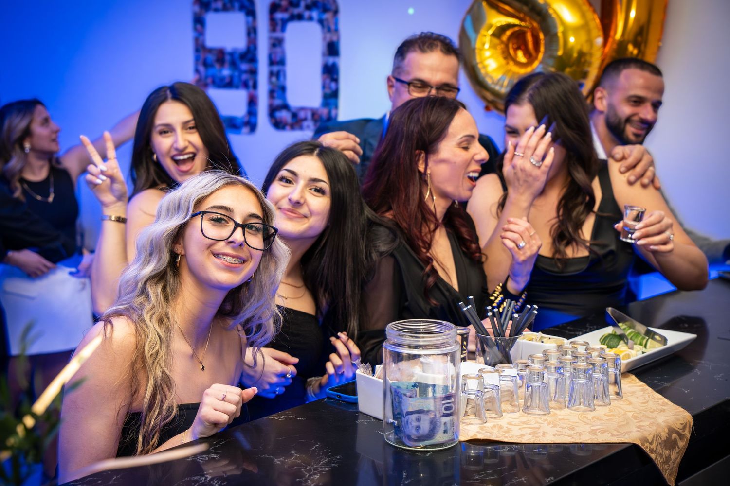 A group enjoying drinks and laughs at a party with gold balloon decorations and blue lighting.