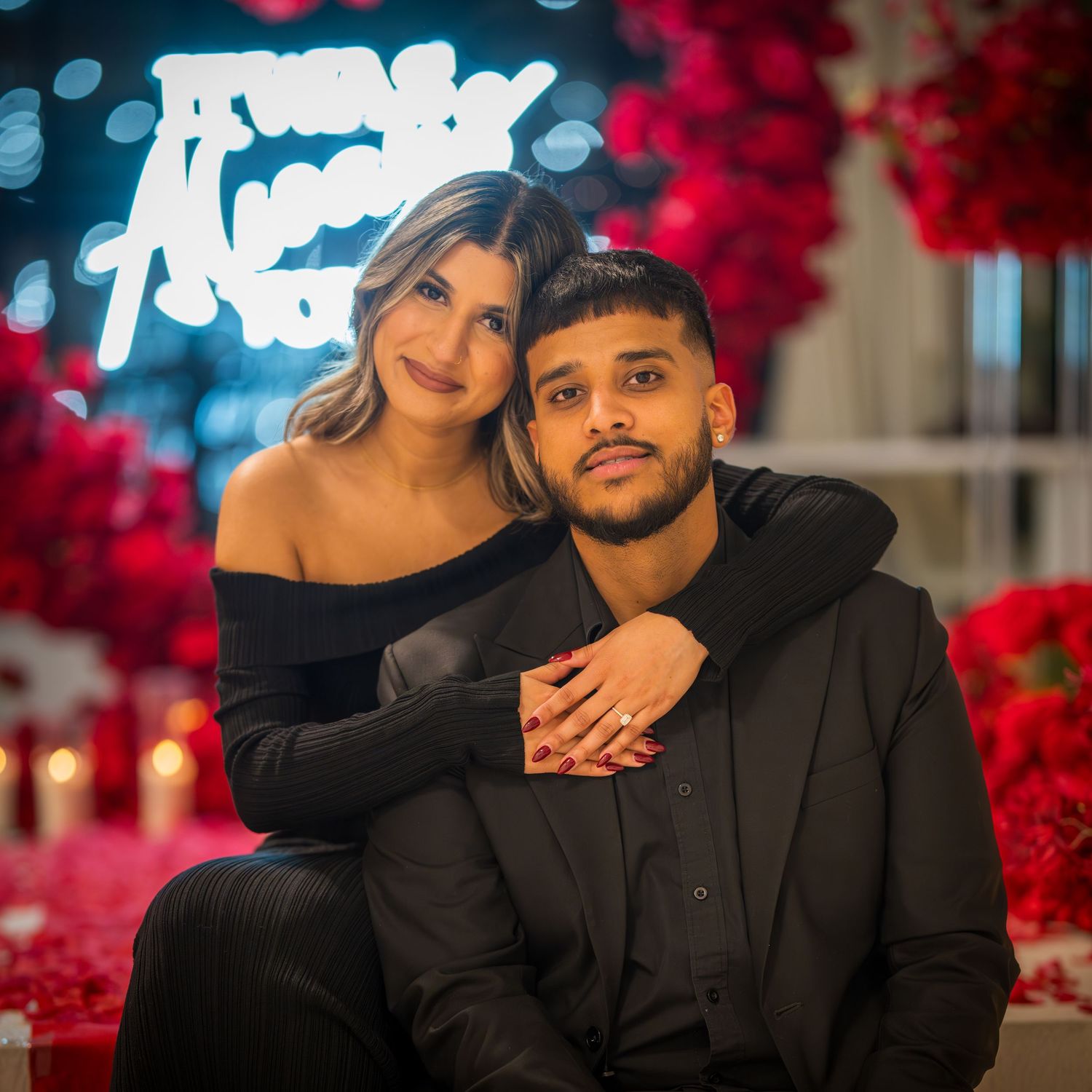 Romantic couple in black attire pose together against a backdrop of vibrant red flowers and blue neon lights.