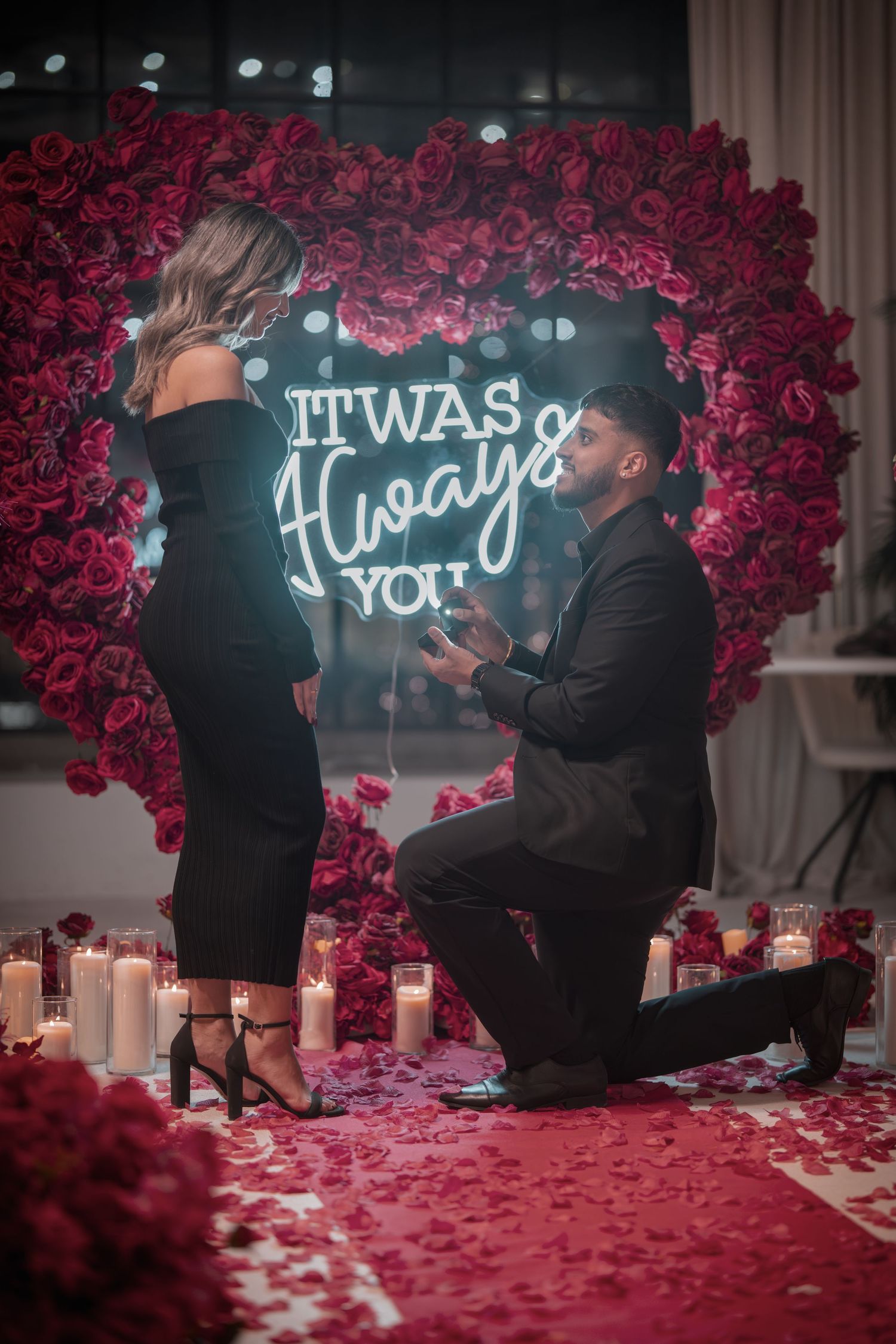 Proposal moment captured with illuminated sign and red rose wall backdrop.