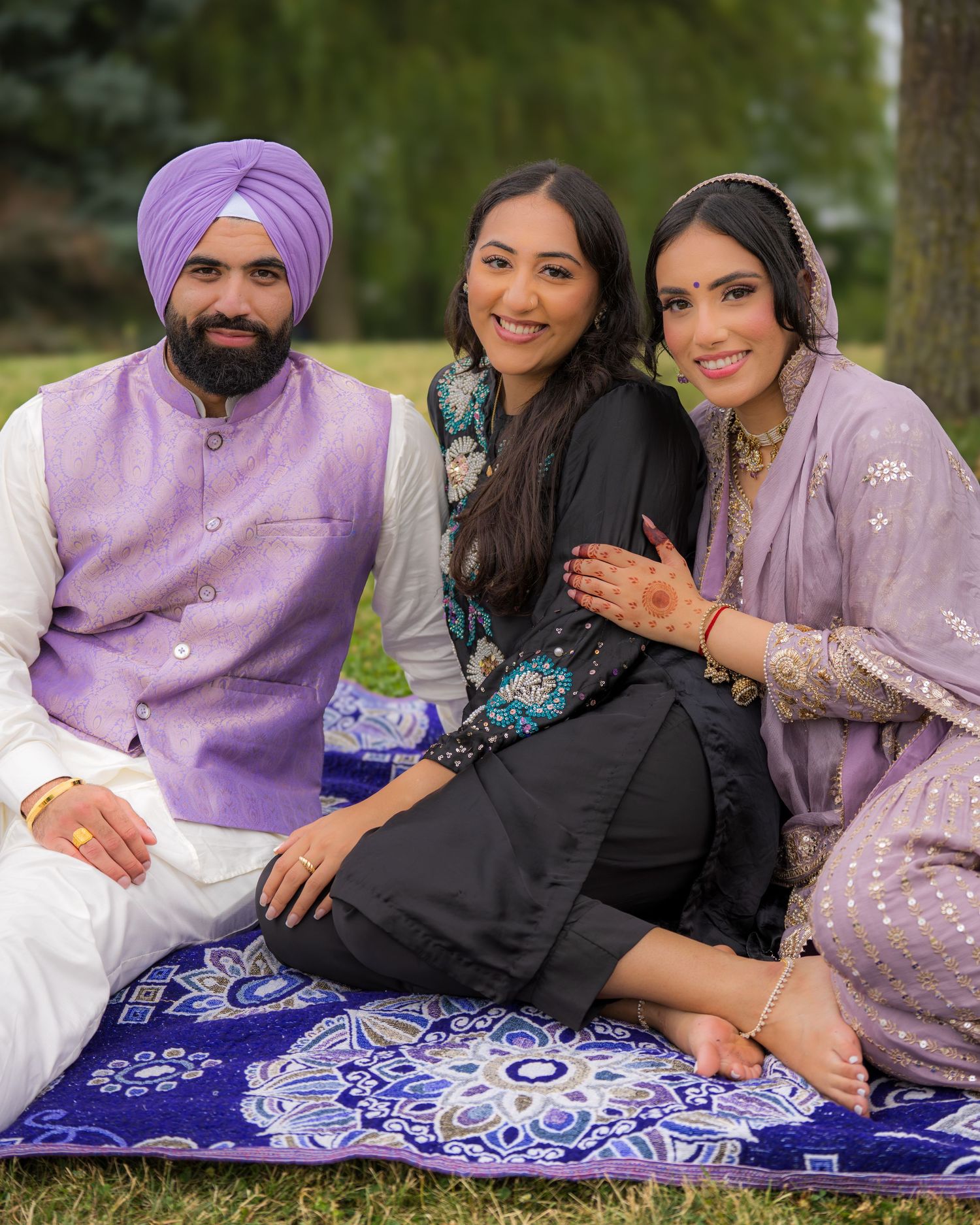 A group poses together on a decorative blue blanket in a park wearing coordinated purple and black traditional outfits.