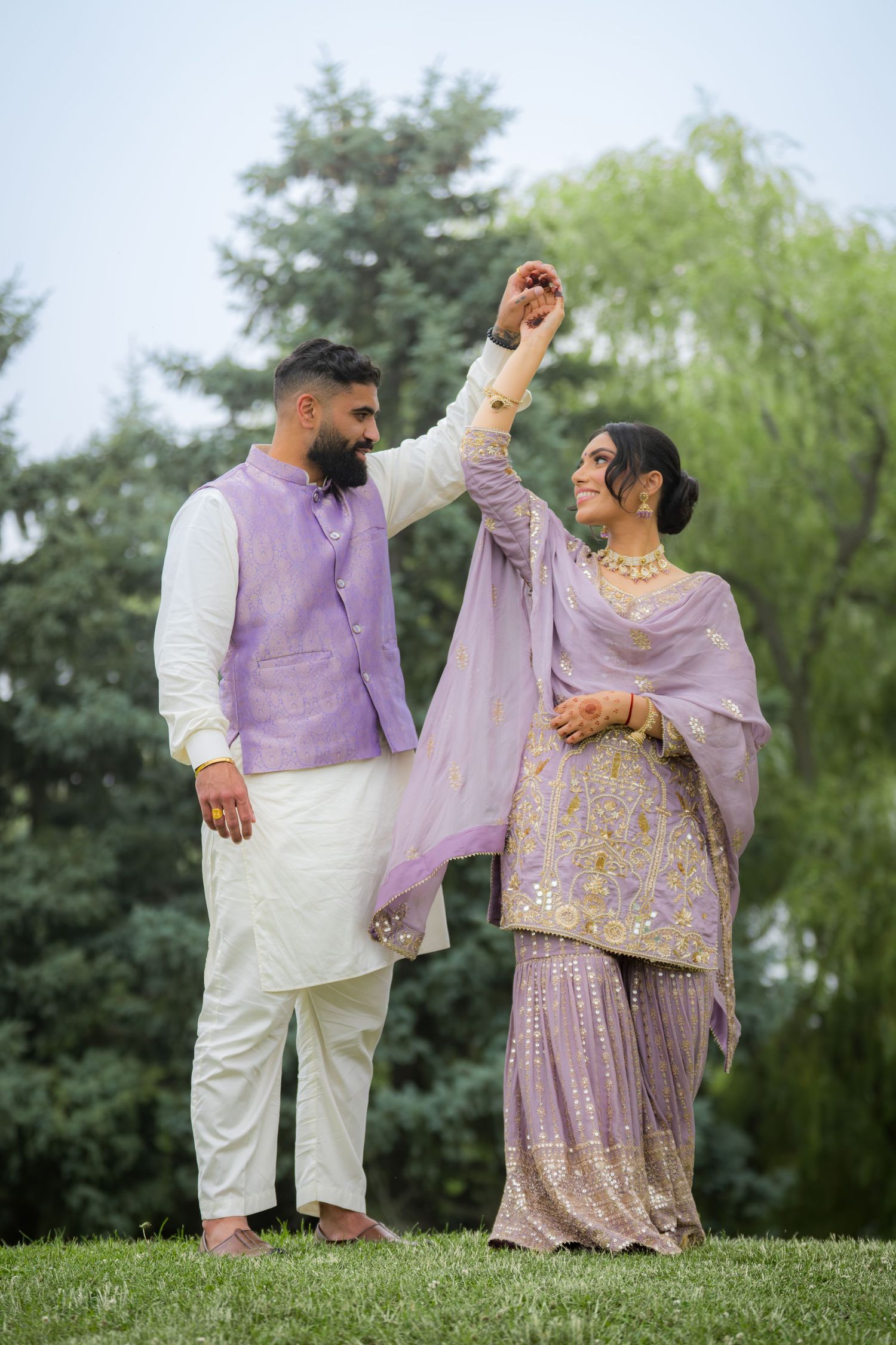 A couple in purple and white traditional dress dance on a grassy hill.