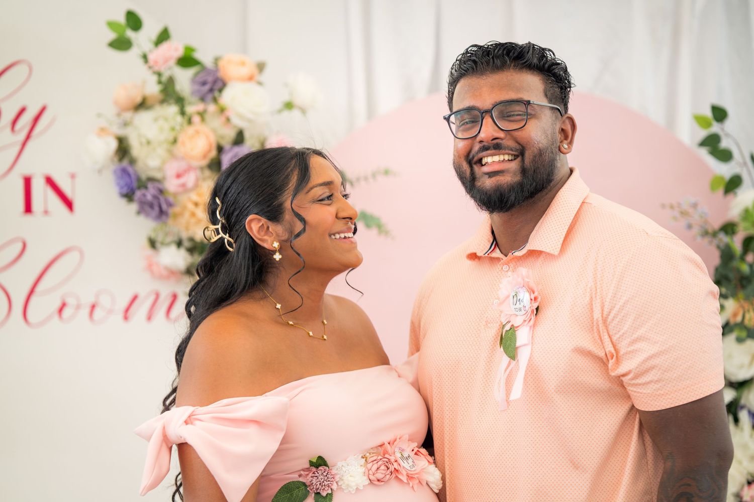 Joyful moment captured at baby shower with couple in matching pink outfits against floral backdrop.