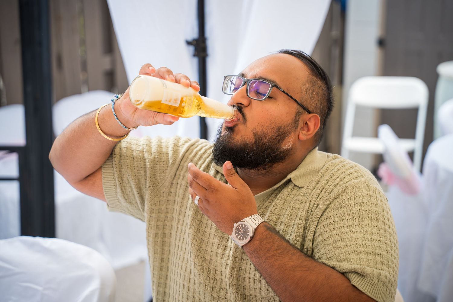 Person in glasses drinking from a glass bottle at a social gathering.