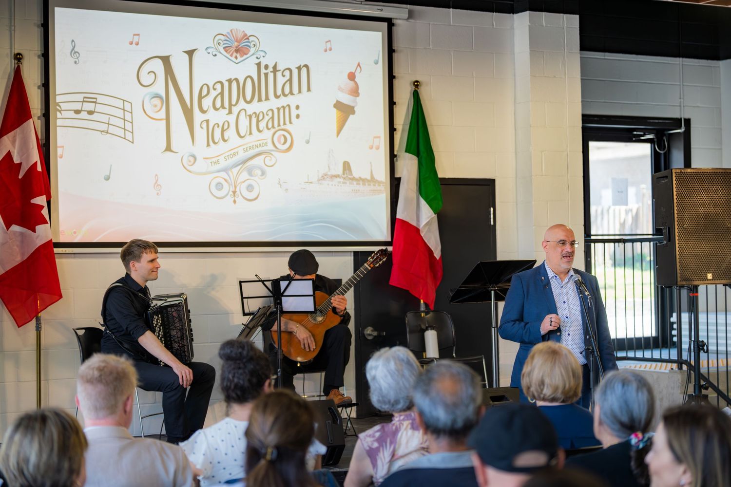 Speaker presenting Neapolitan Ice Cream event with musicians and Canadian and Italian flags on stage.