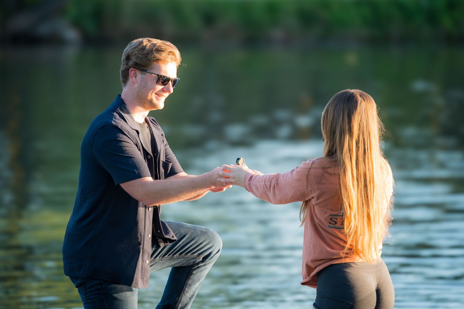 Man kneeling on one knee proposing to woman by a lake in golden hour light.