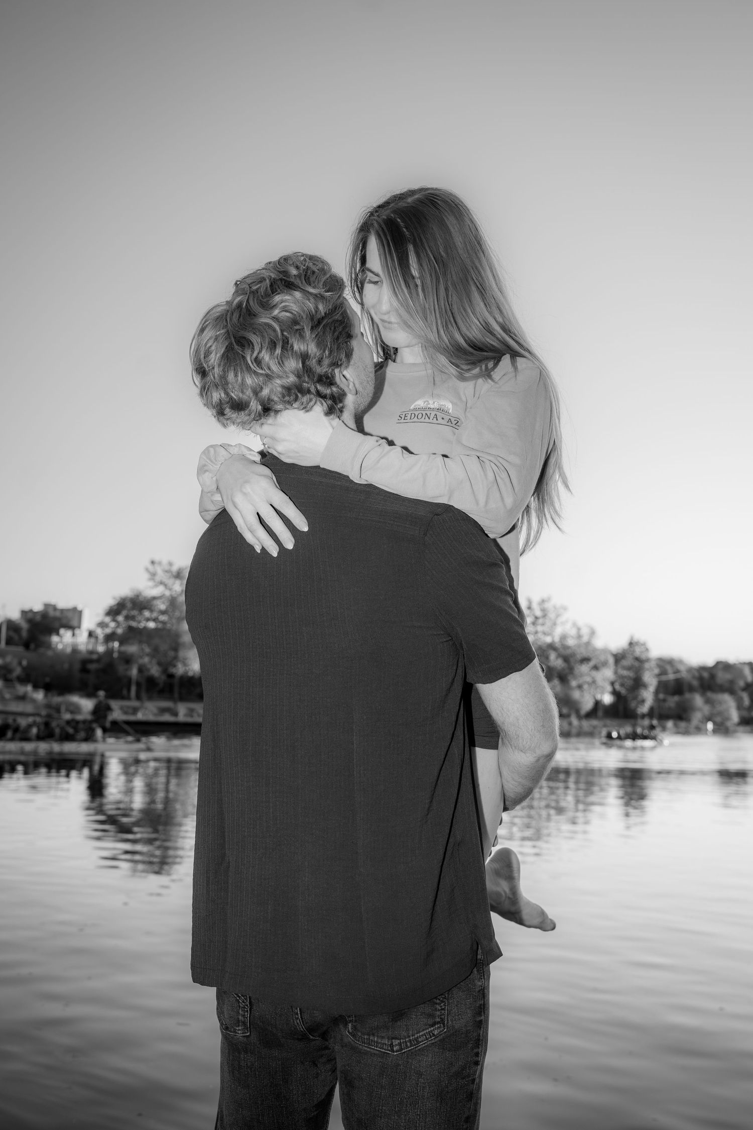 Black and white photo of a couple embracing by a lake at sunset.