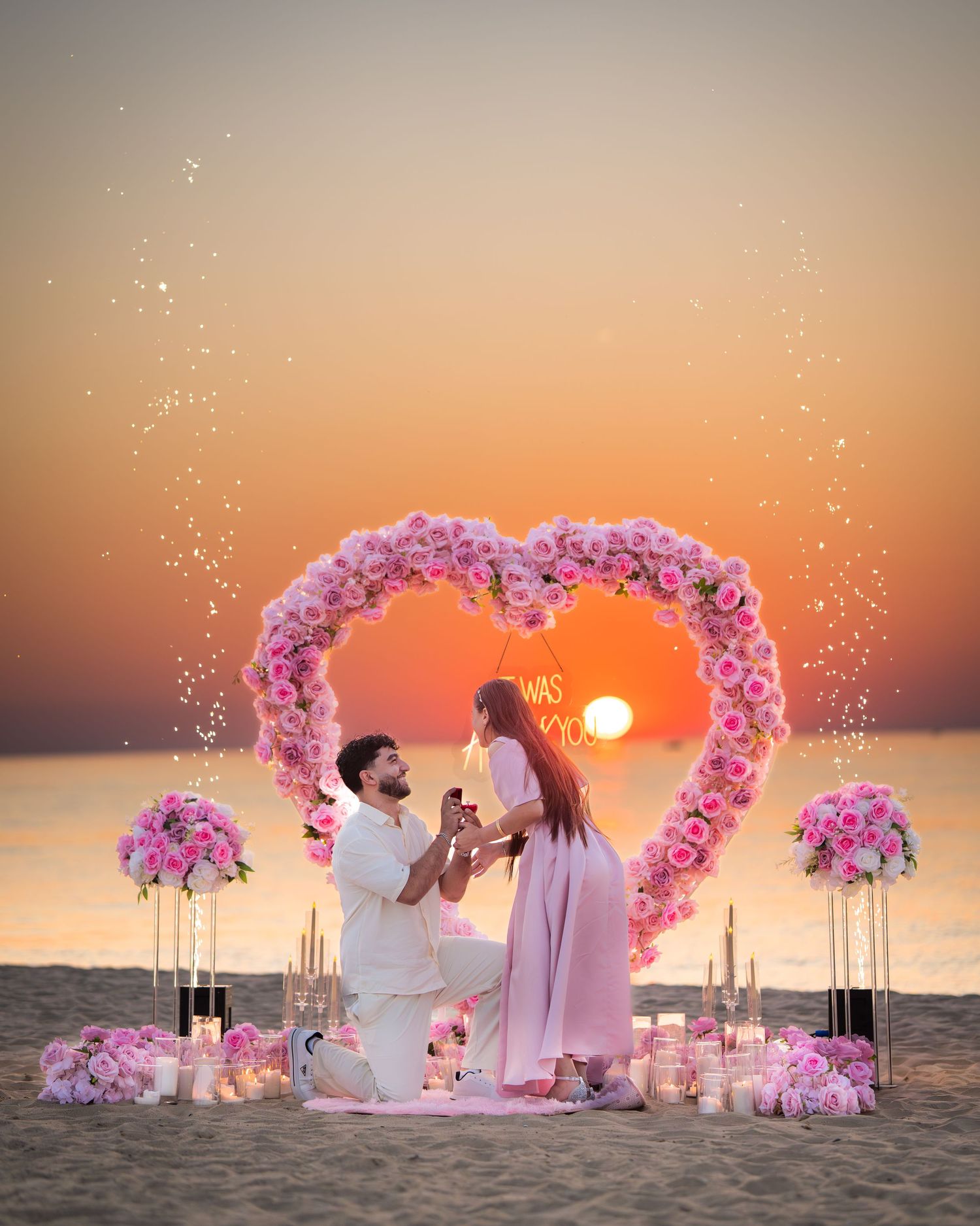 A romantic beach proposal setup with a pink floral heart arch and candles against a stunning sunset backdrop.