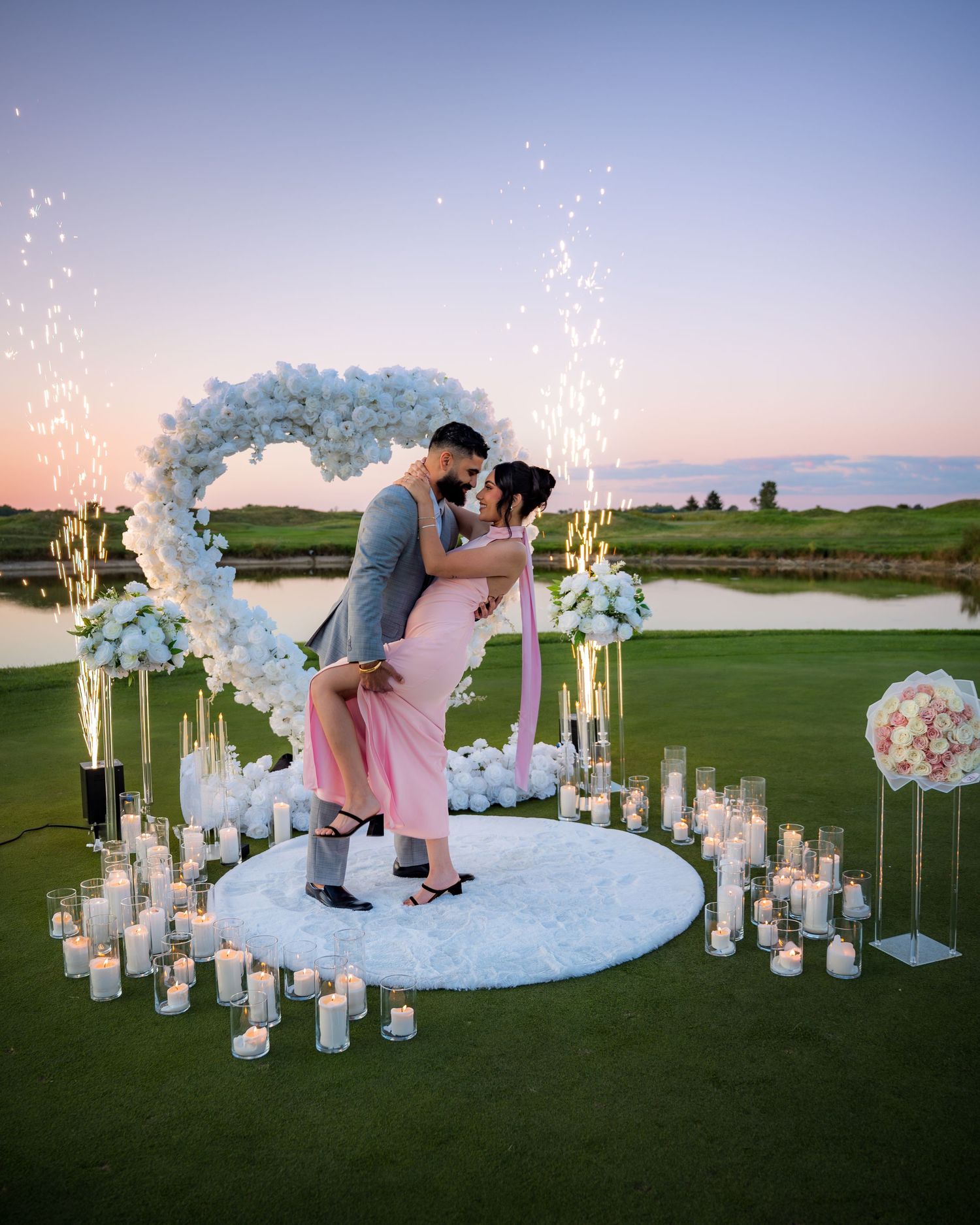 Couple sharing romantic moment under floral arch with sparkling lights and candles on golf course.
