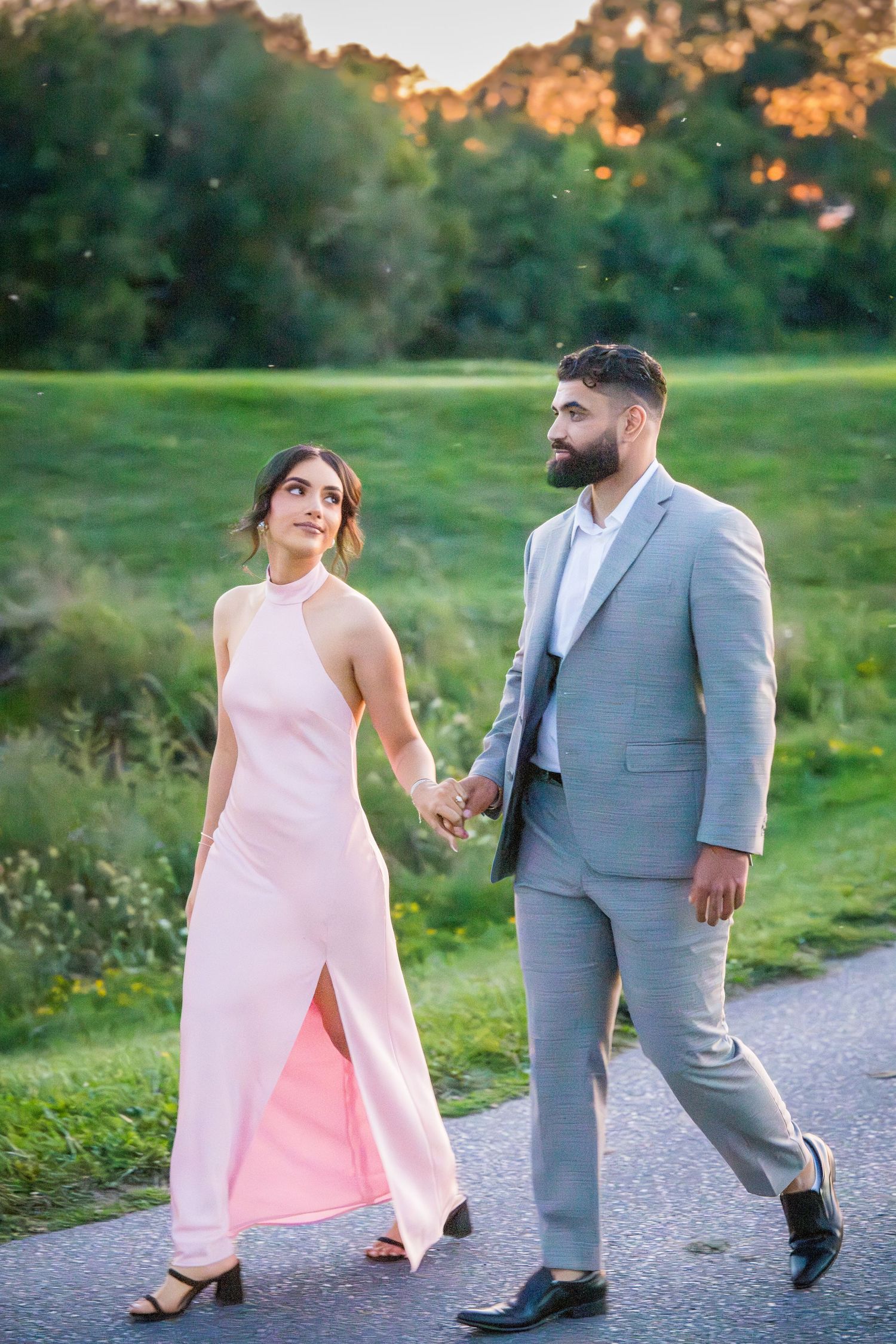 Couple strolling on gravel path at sunset, wearing pink dress and gray suit.