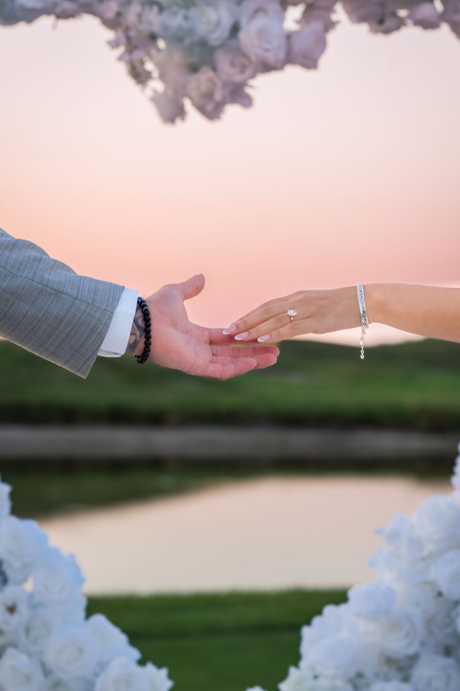 Close up of hands reaching towards each other with white floral arch and pink sky.