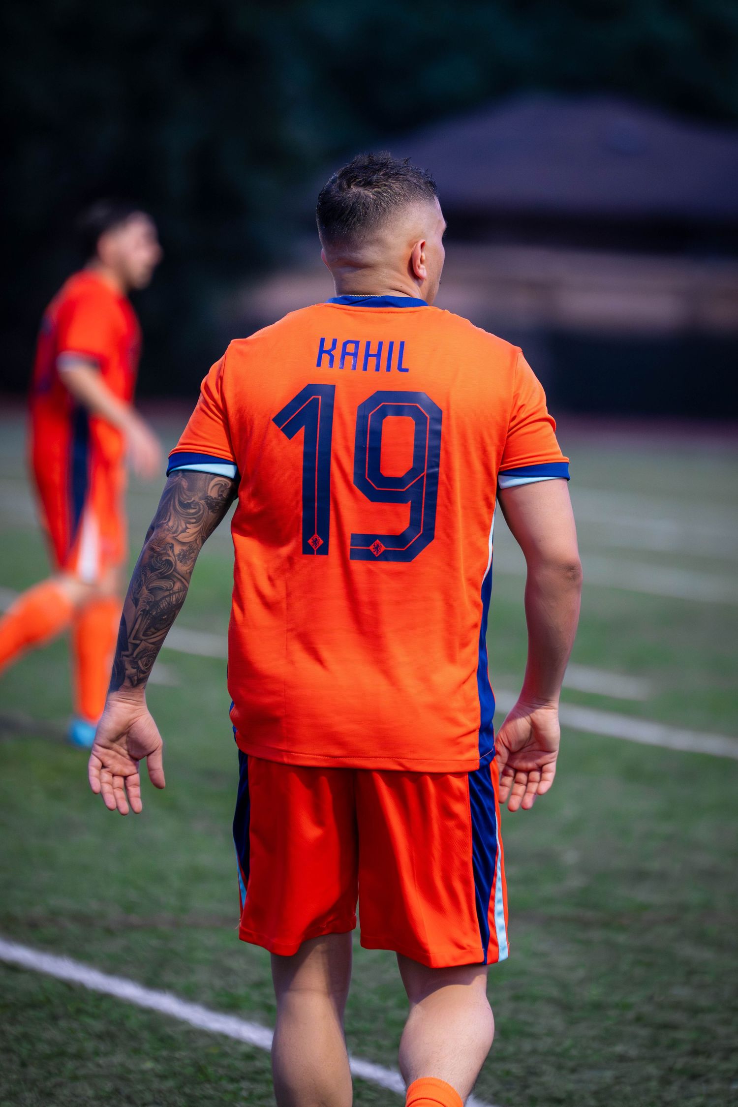 Soccer player wearing orange jersey number 19 with 'RAHIL' text stretches on grass field during match.