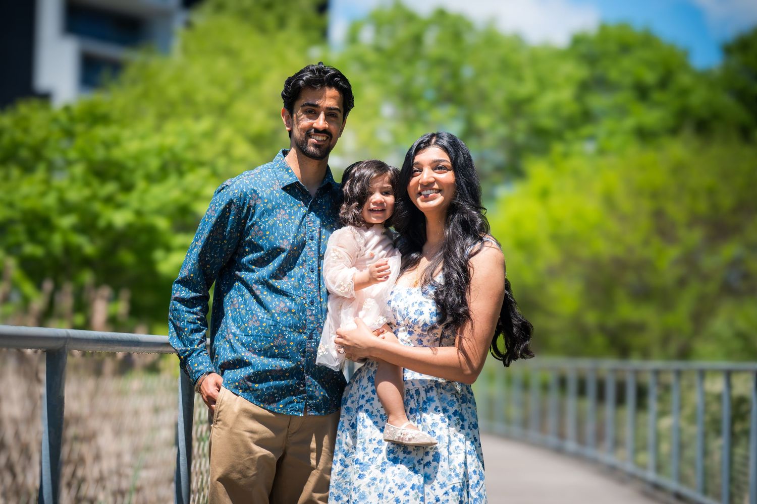 A smiling family of three poses on a bridge, with parents holding their young daughter outdoors.