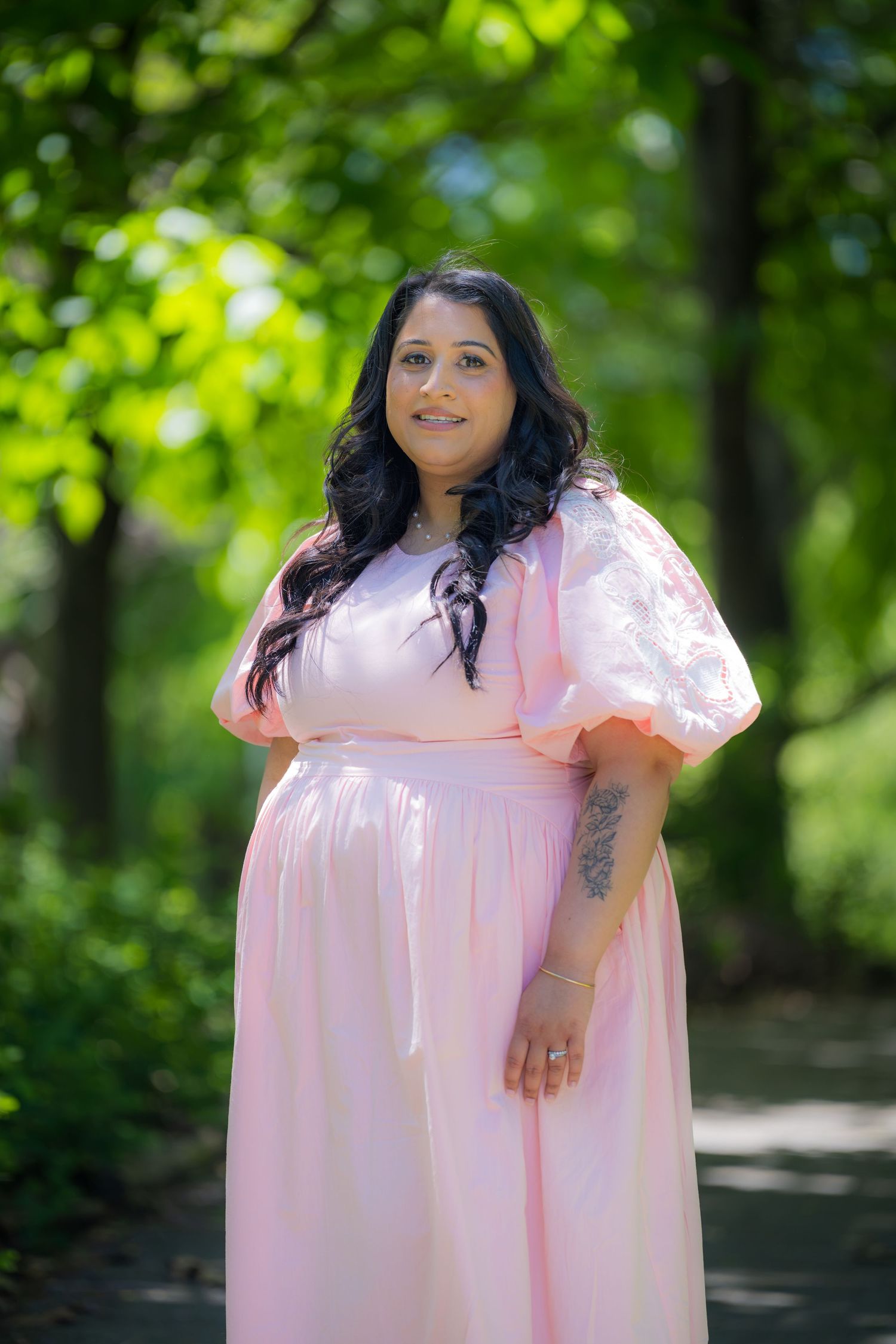 Smiling woman in pink puff sleeve maxi dress standing on tree-lined garden path.