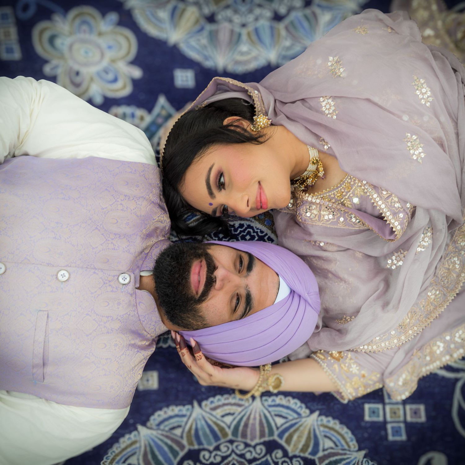 A couple in traditional Punjabi attire lying on decorative cushions against a blue patterned background.