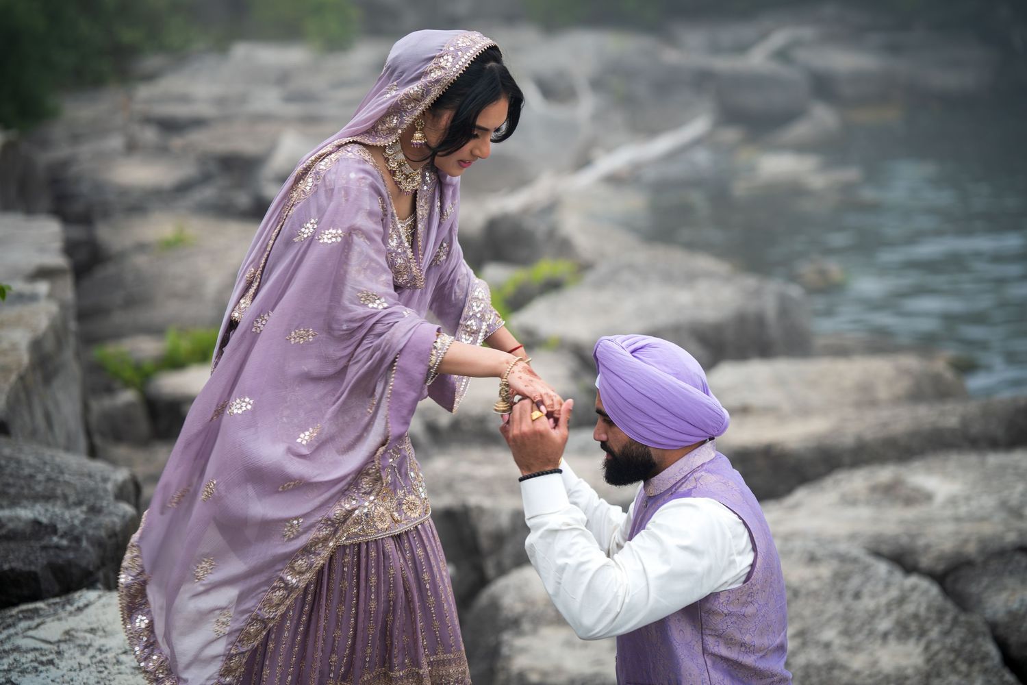 Romantic couple in traditional Punjabi attire pose together on rocky shore.