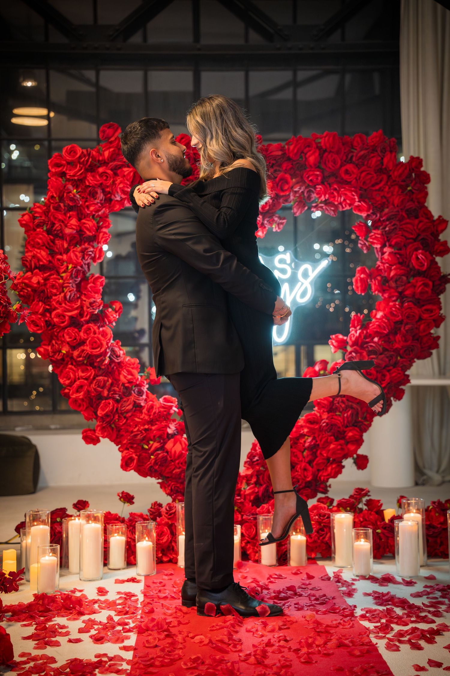 Couple embraces in romantic setting with red rose walls and candlelit floor.