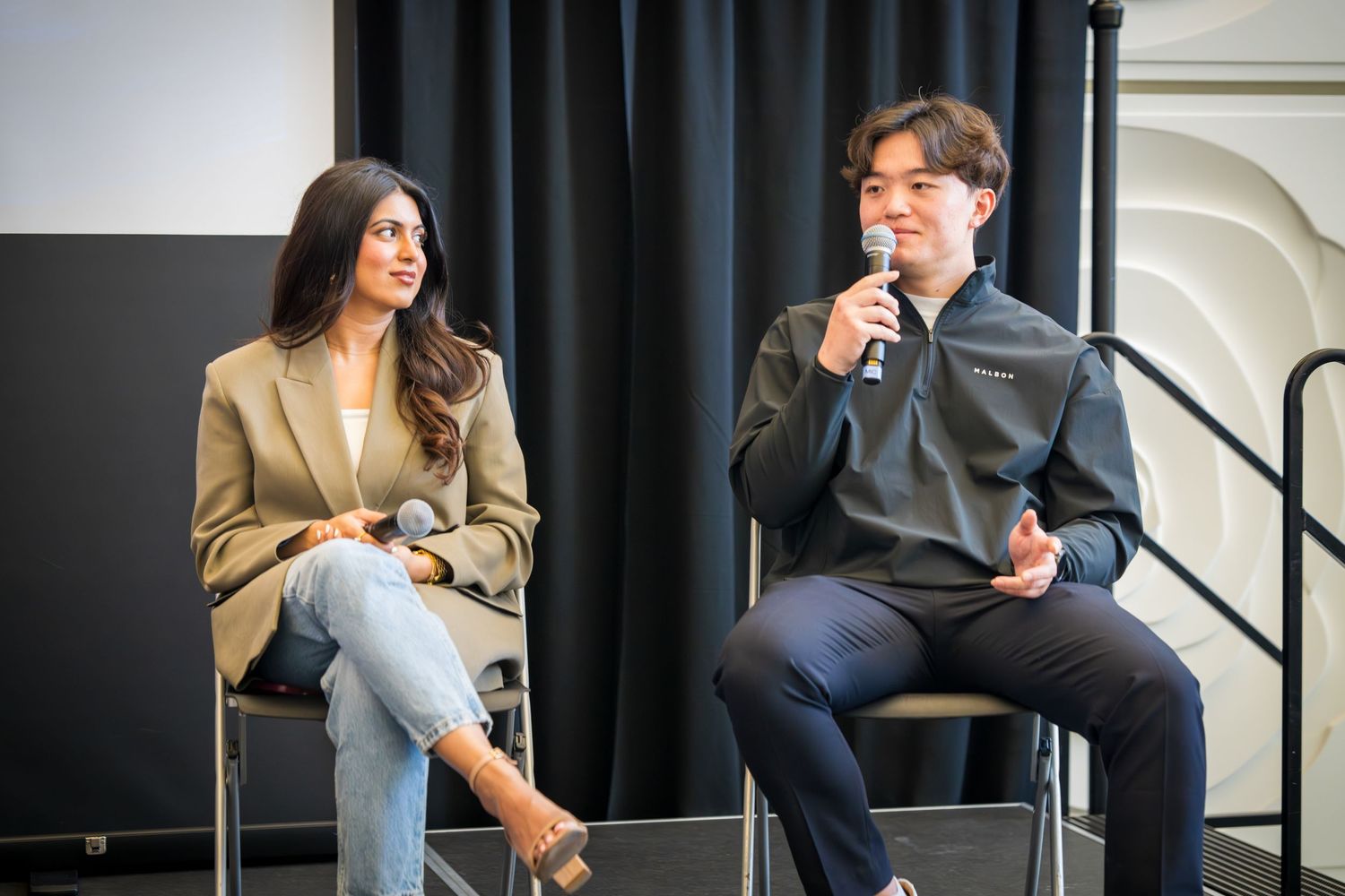 Panel discussion participants seated in chairs on stage with black curtain backdrop and white textured wall.