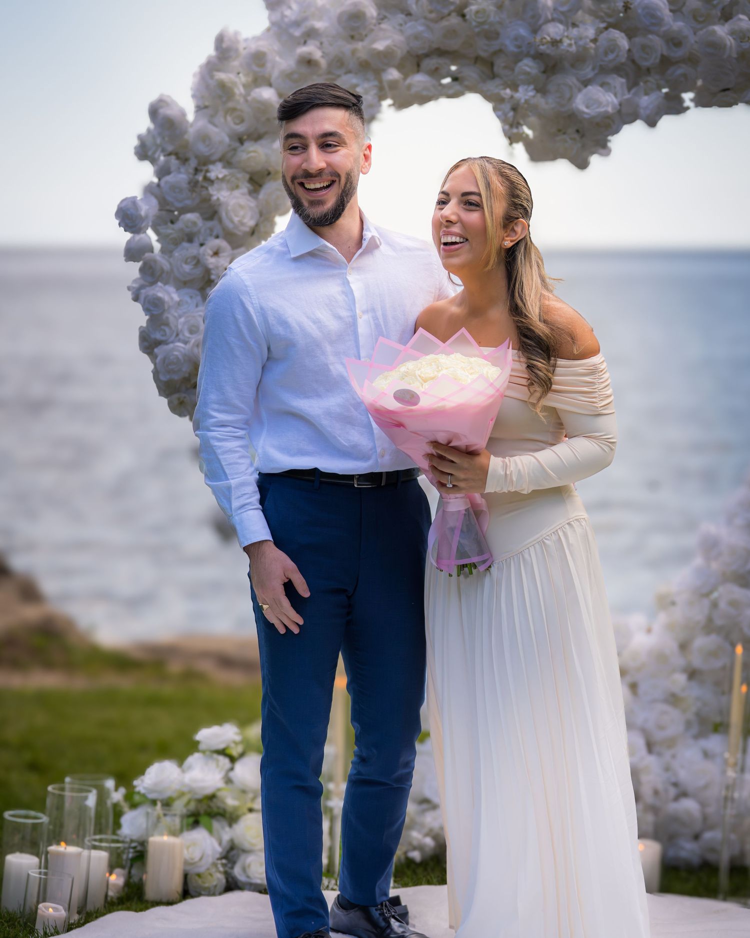 Joyful moment with pink bouquet under white balloon arch at waterfront ceremony.