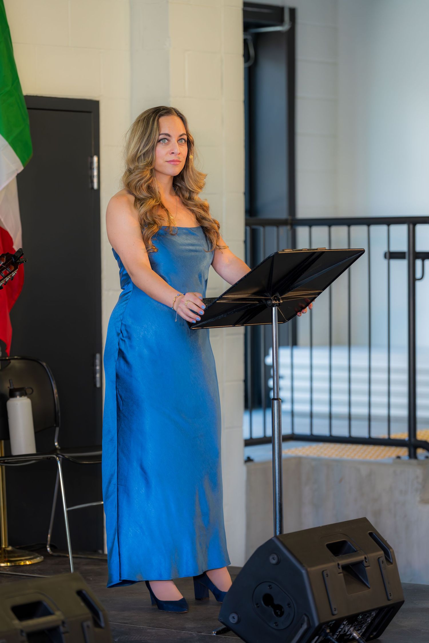 Woman in blue satin gown presenting at podium with music stand at formal Italian event.