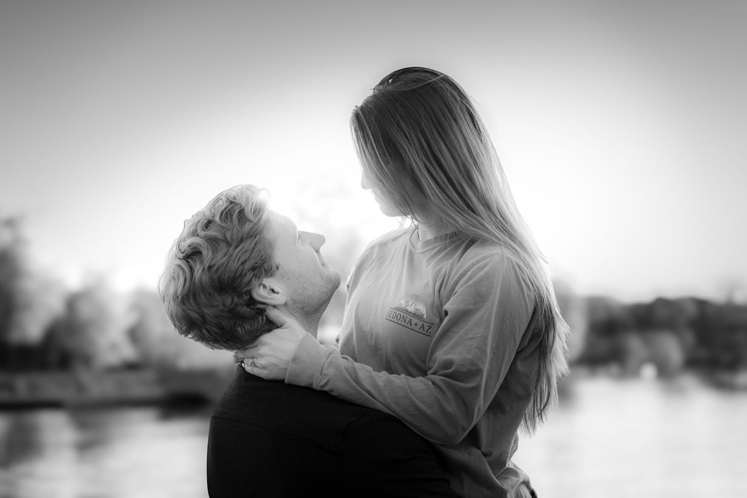 Black and white romantic photo of a couple embracing and almost kissing by a sunlit lake.